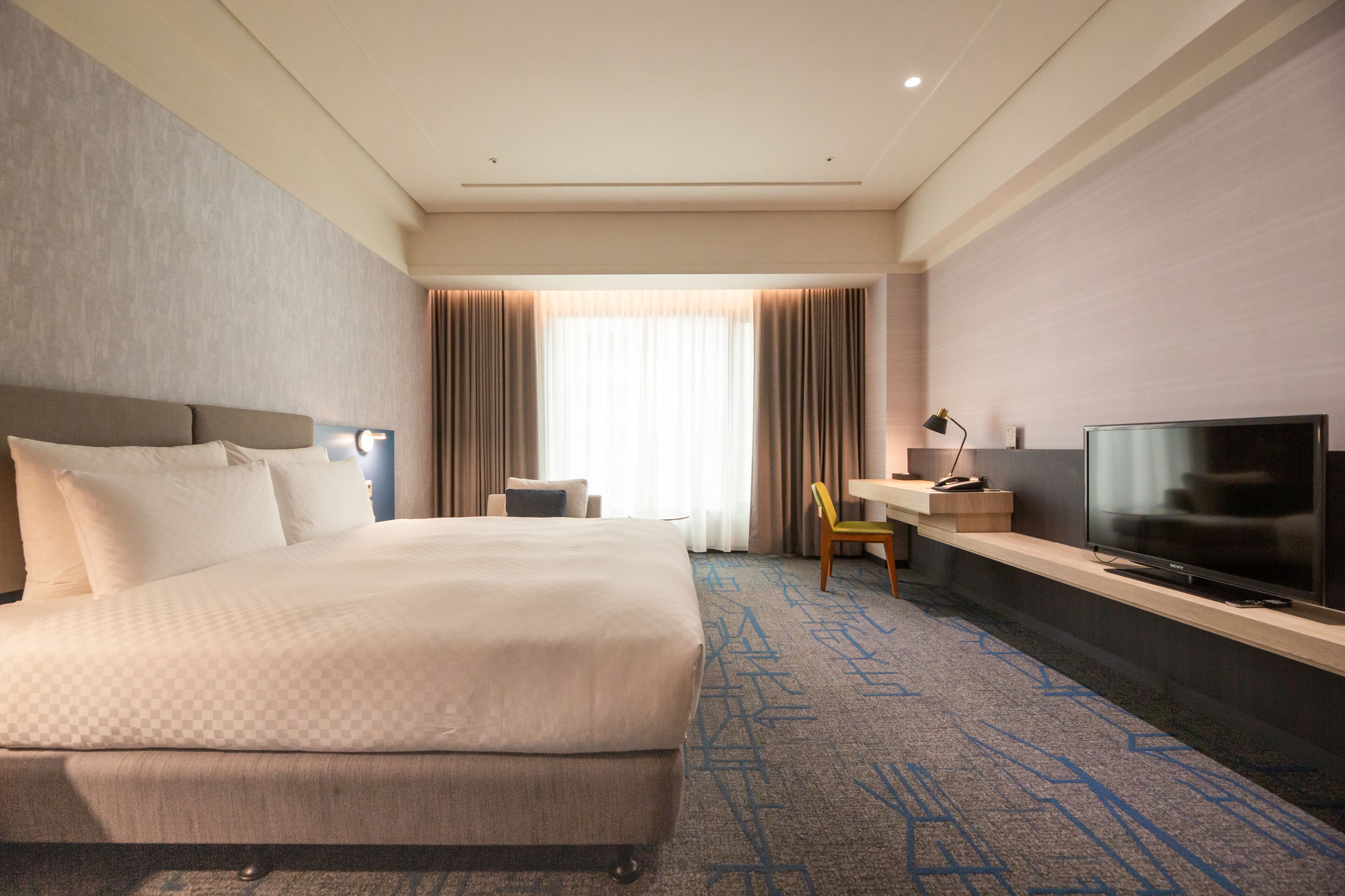 A hotel room with a large bed, a flat-screen TV, a desk with a chair, and a window with curtains.