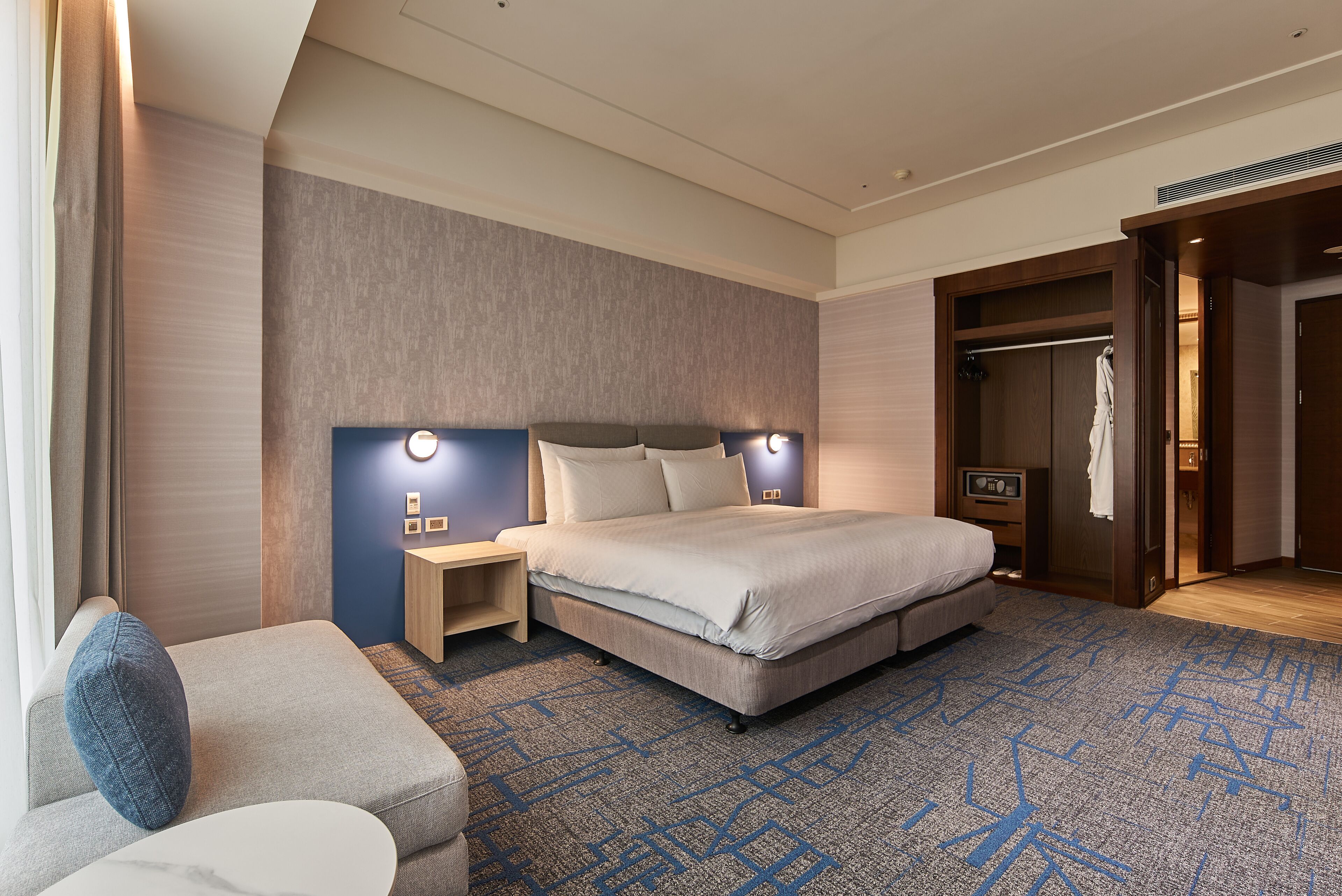 A modern hotel room with a large bed, a bedside table, a nightstand, a seating area with a sofa, and a walk-in closet.