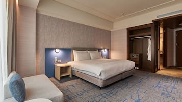 A modern hotel room with a large bed, a bedside table, a nightstand, a seating area with a sofa, and a walk-in closet.