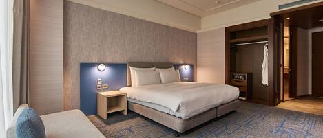A modern hotel room with a large bed, a bedside table, a nightstand, a seating area with a sofa, and a walk-in closet.