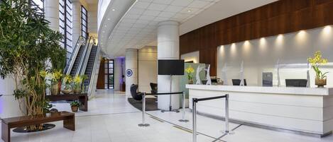 A modern hotel lobby with a reception desk, seating area, and an escalator.