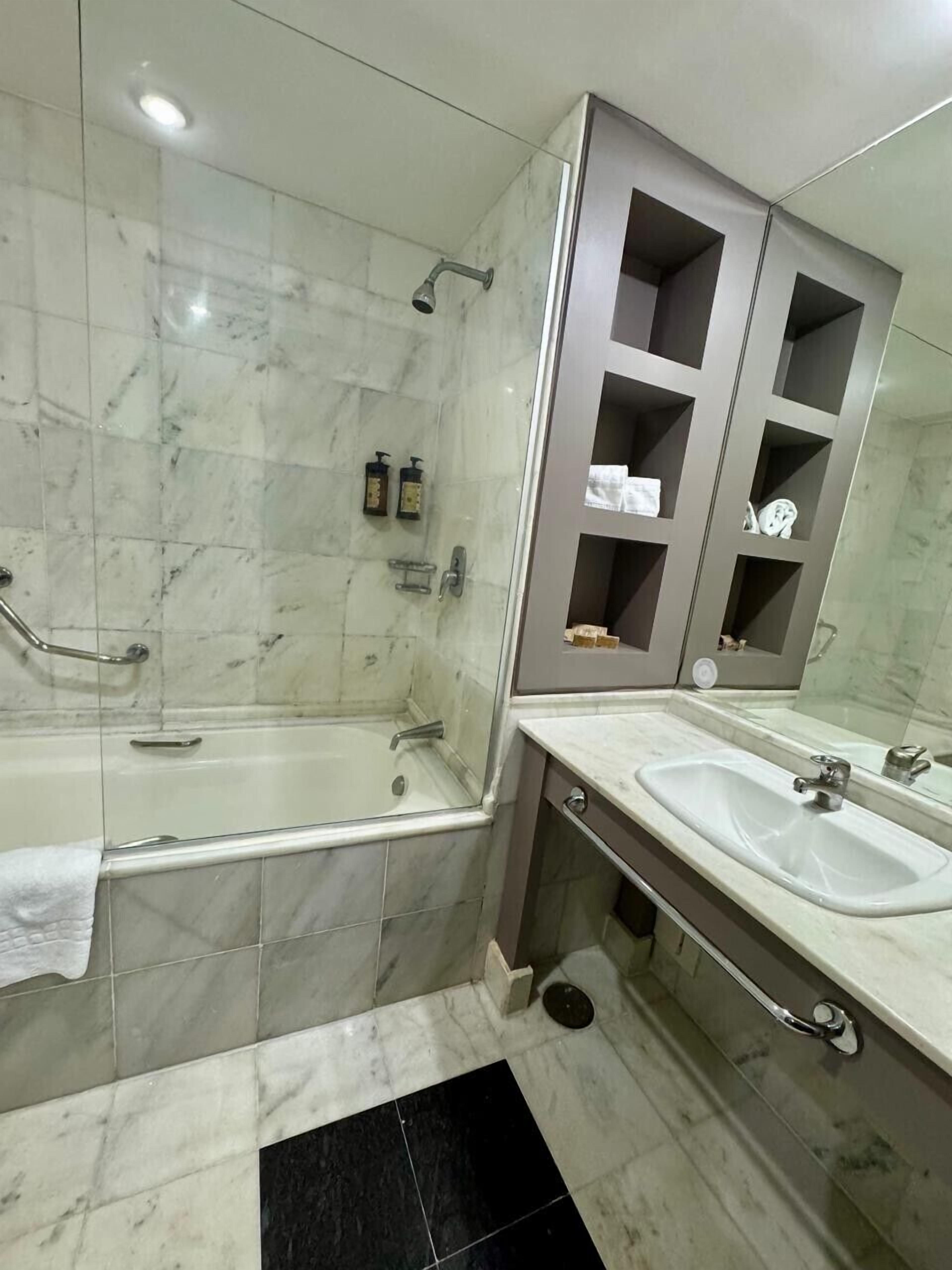 Een moderne badkamer met een badkuip, twee wastafels, een glazen douchecabine en een badkamermeubel met planken.