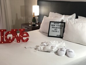 A hotel room with a bed, a nightstand with a bottle and glasses, a sign with text, and slippers.