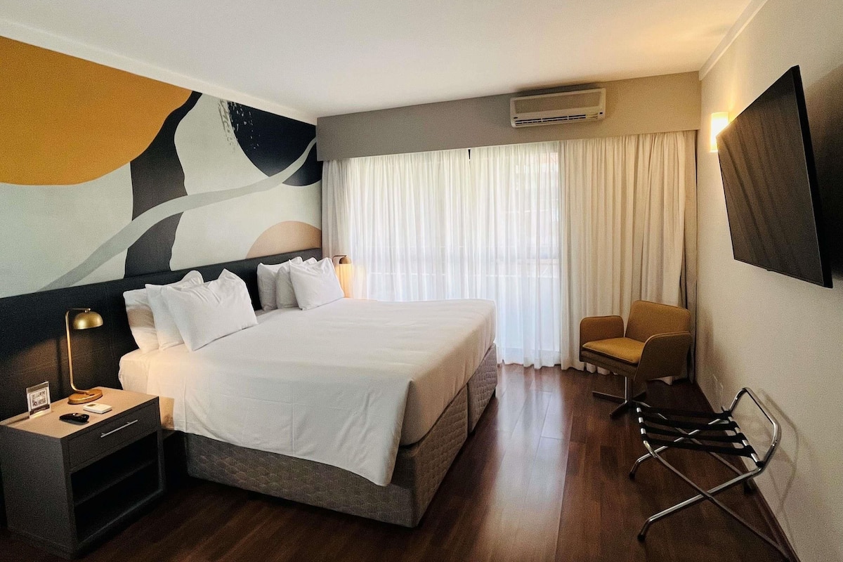 A hotel room with a bed, a chair, a desk, a TV, and an air conditioning unit.
