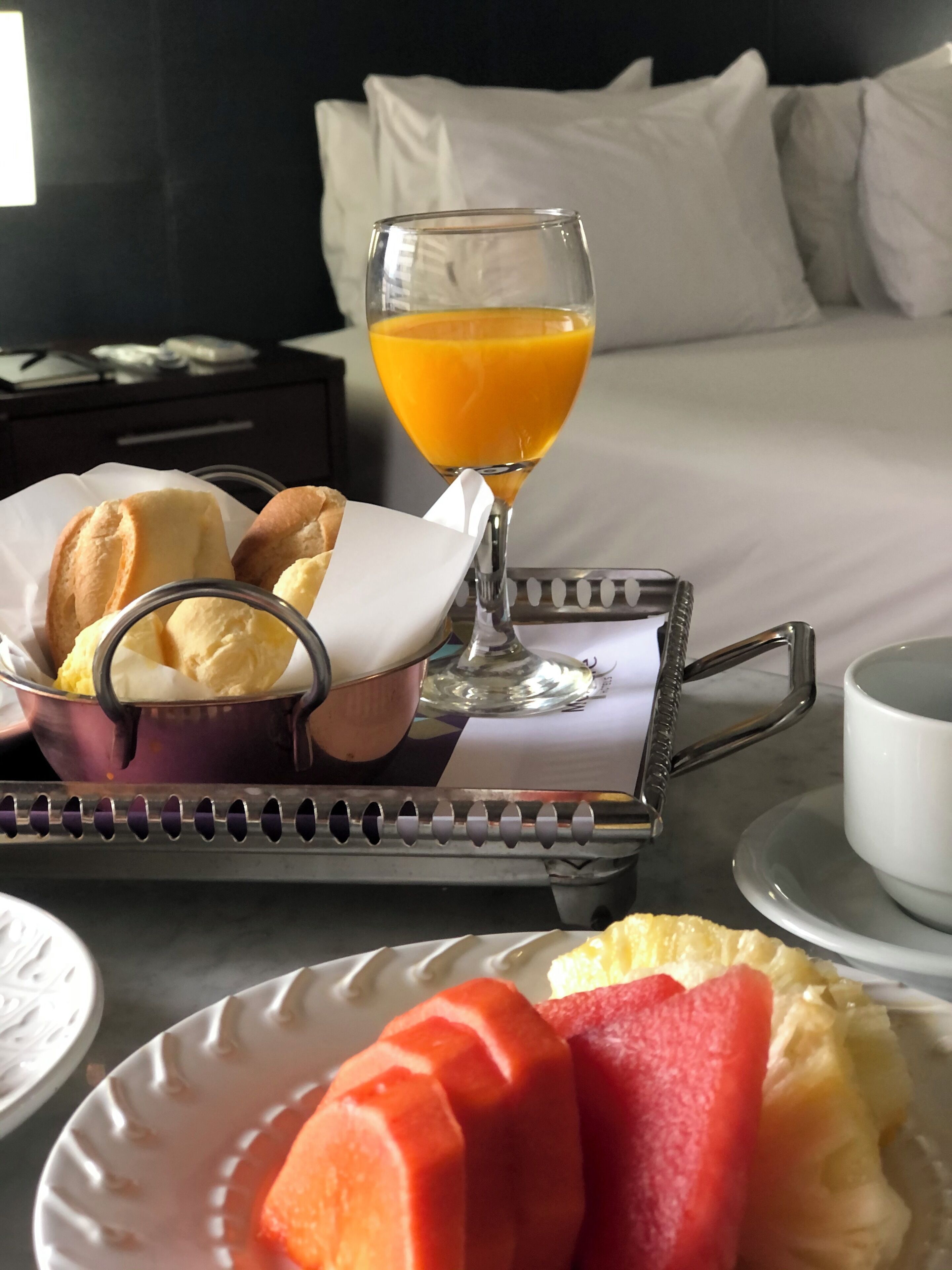 Bandeja de café da manhã com pão, manteiga e um copo de suco de laranja sobre uma mesa de quarto de hotel.