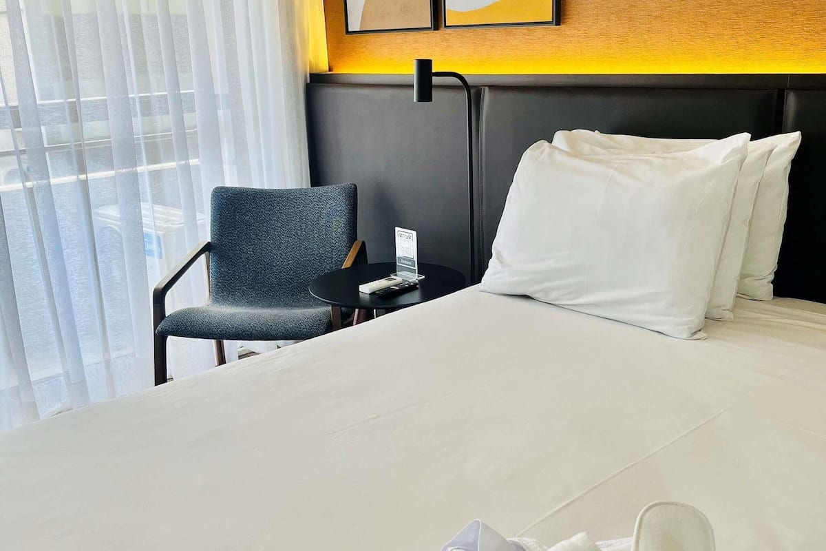Hotellihuone, jossa on sänky, tuoli, pieni pöytä ja kannettava tietokone.