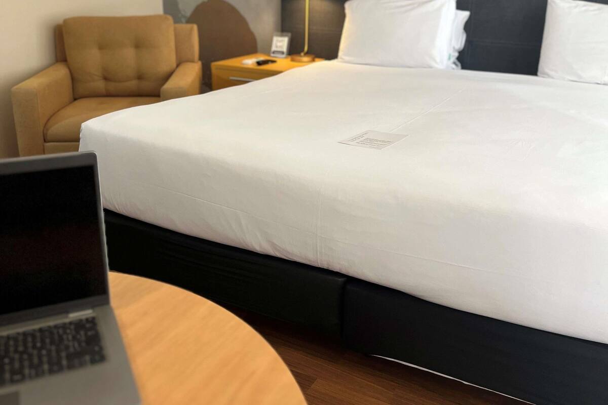 A hotel room with a bed, a desk with a laptop, a chair, a nightstand, and a lamp.