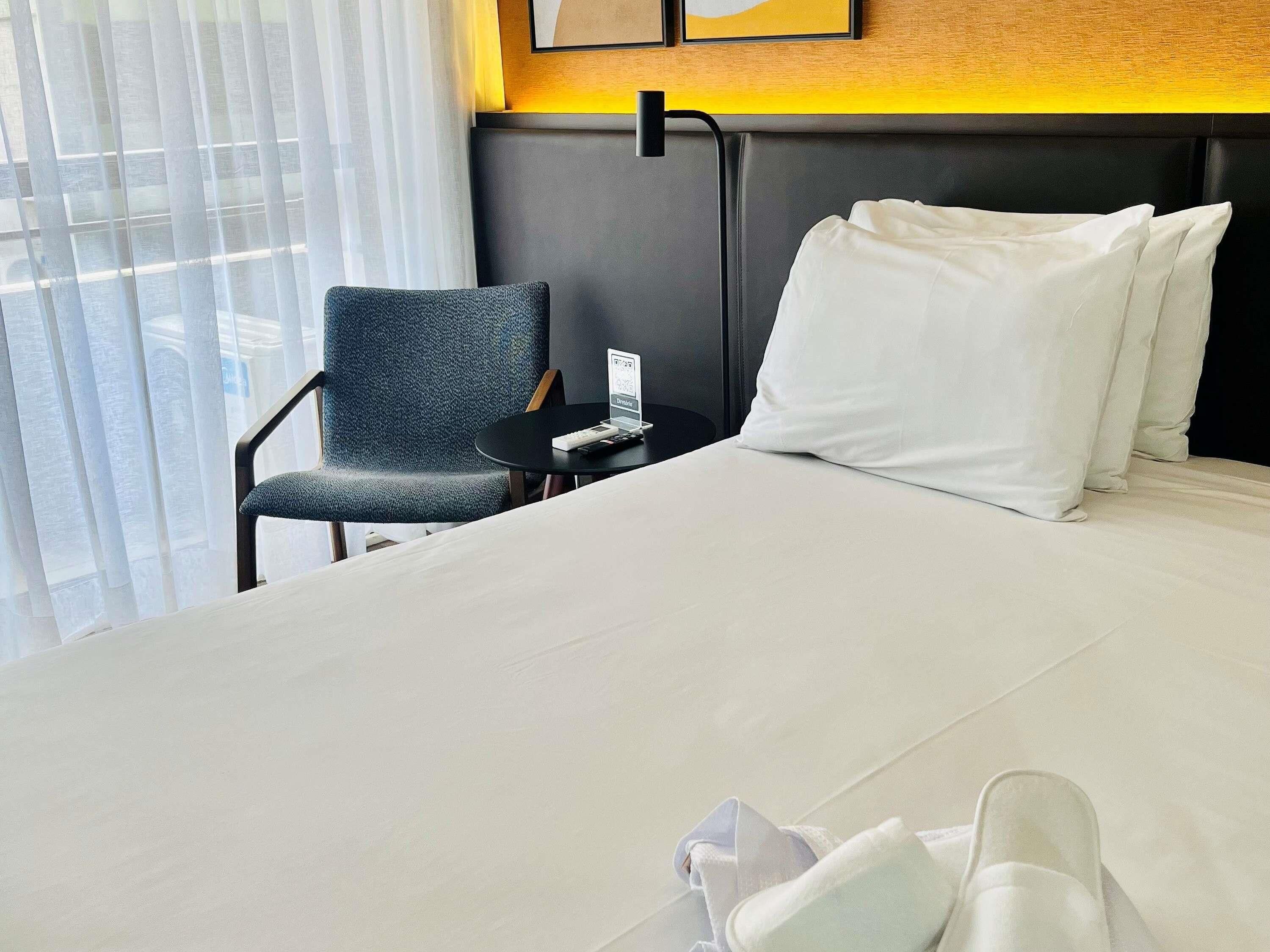 A hotel room with a bed, a chair, a small table with a remote control, and a window with curtains.