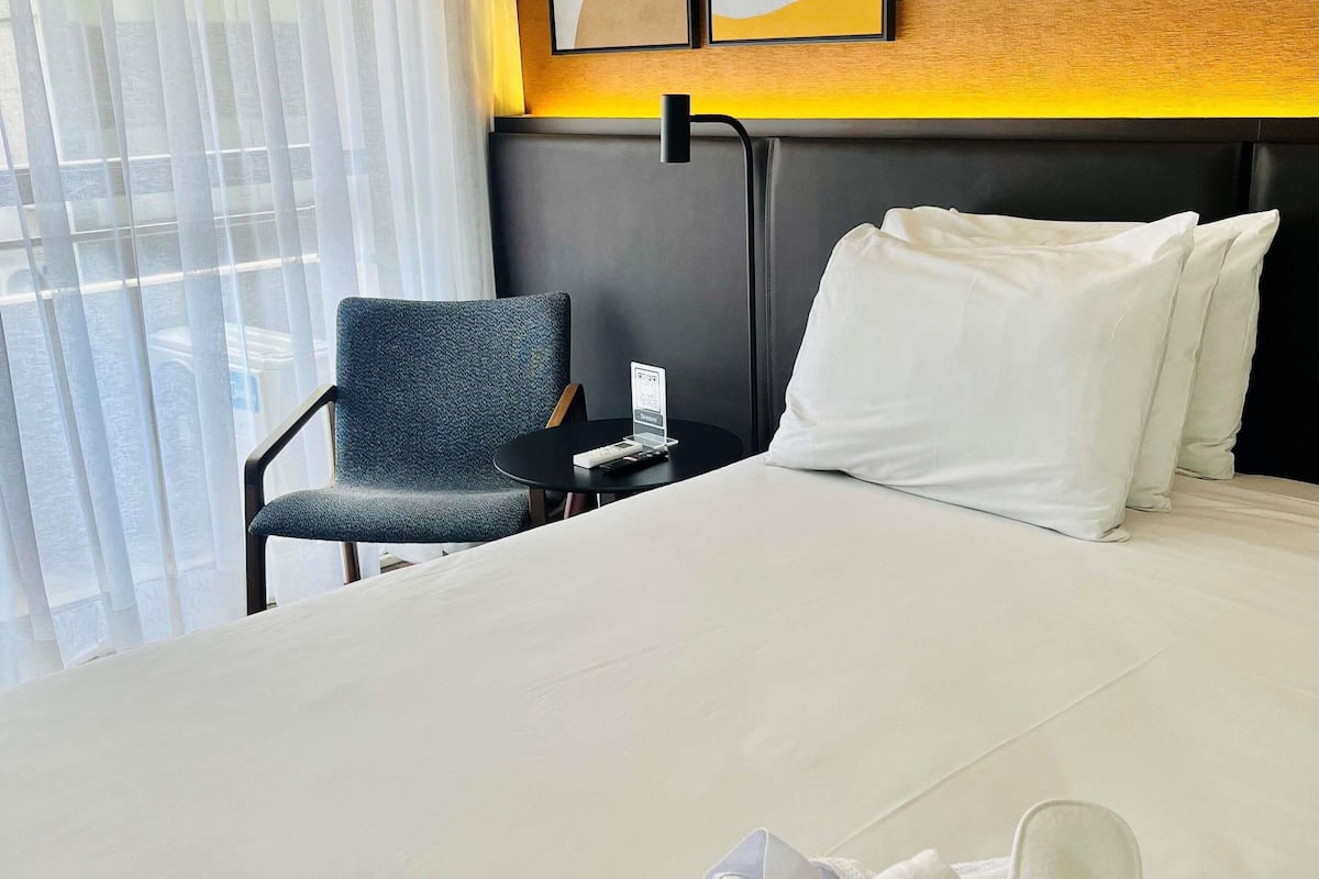 A hotel room with a bed, a chair, a small table with a remote control, and a window with curtains.
