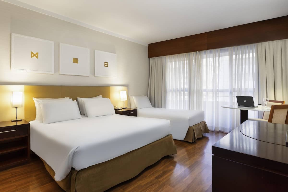 A hotel room with two beds, a desk with a laptop, and a large window with curtains.