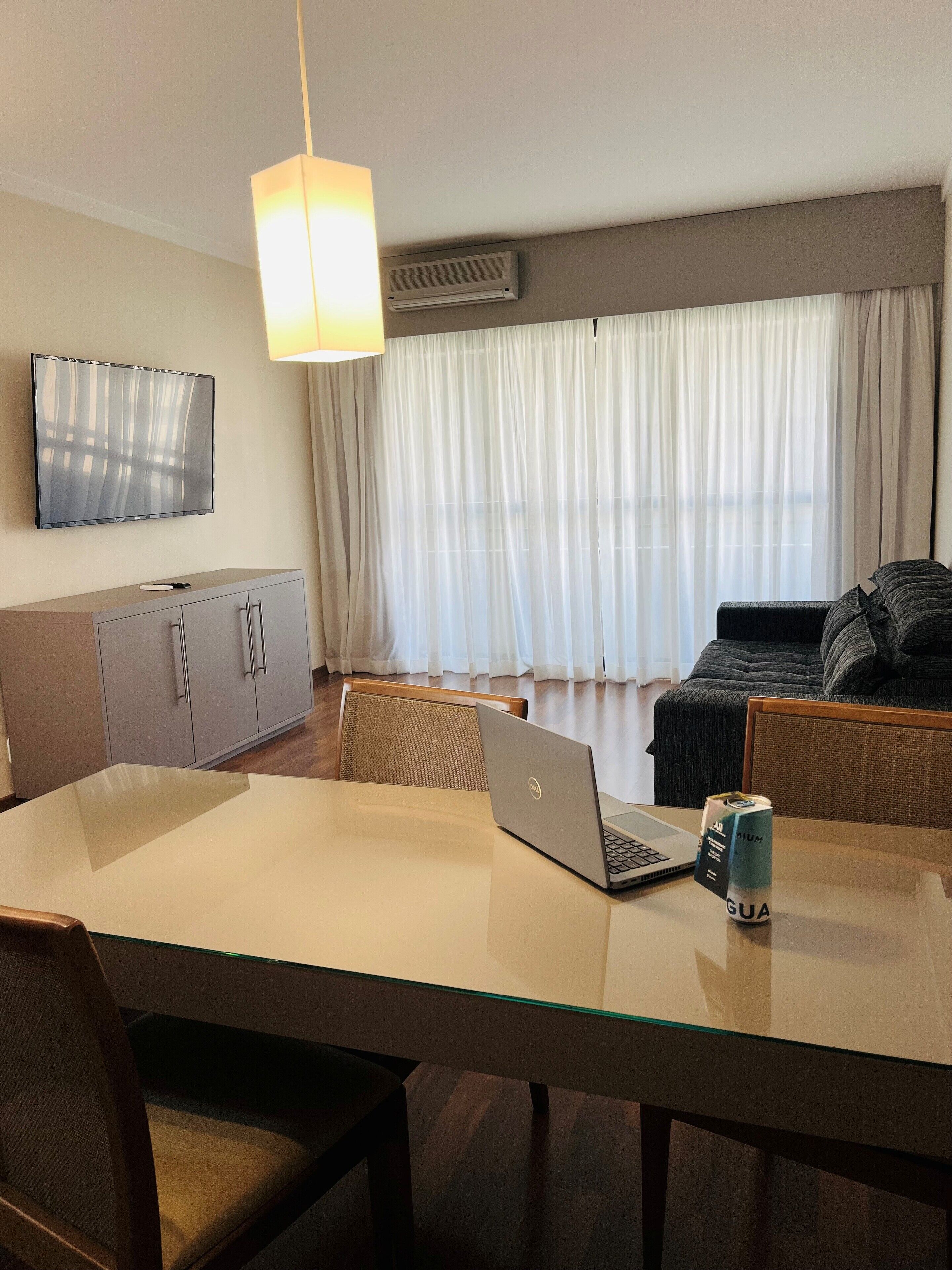 Una sala de estar moderna con una mesa de vidrio para comer, una computadora portátil y una lata sobre la mesa. Hay un sofá y un televisor montado en la pared.