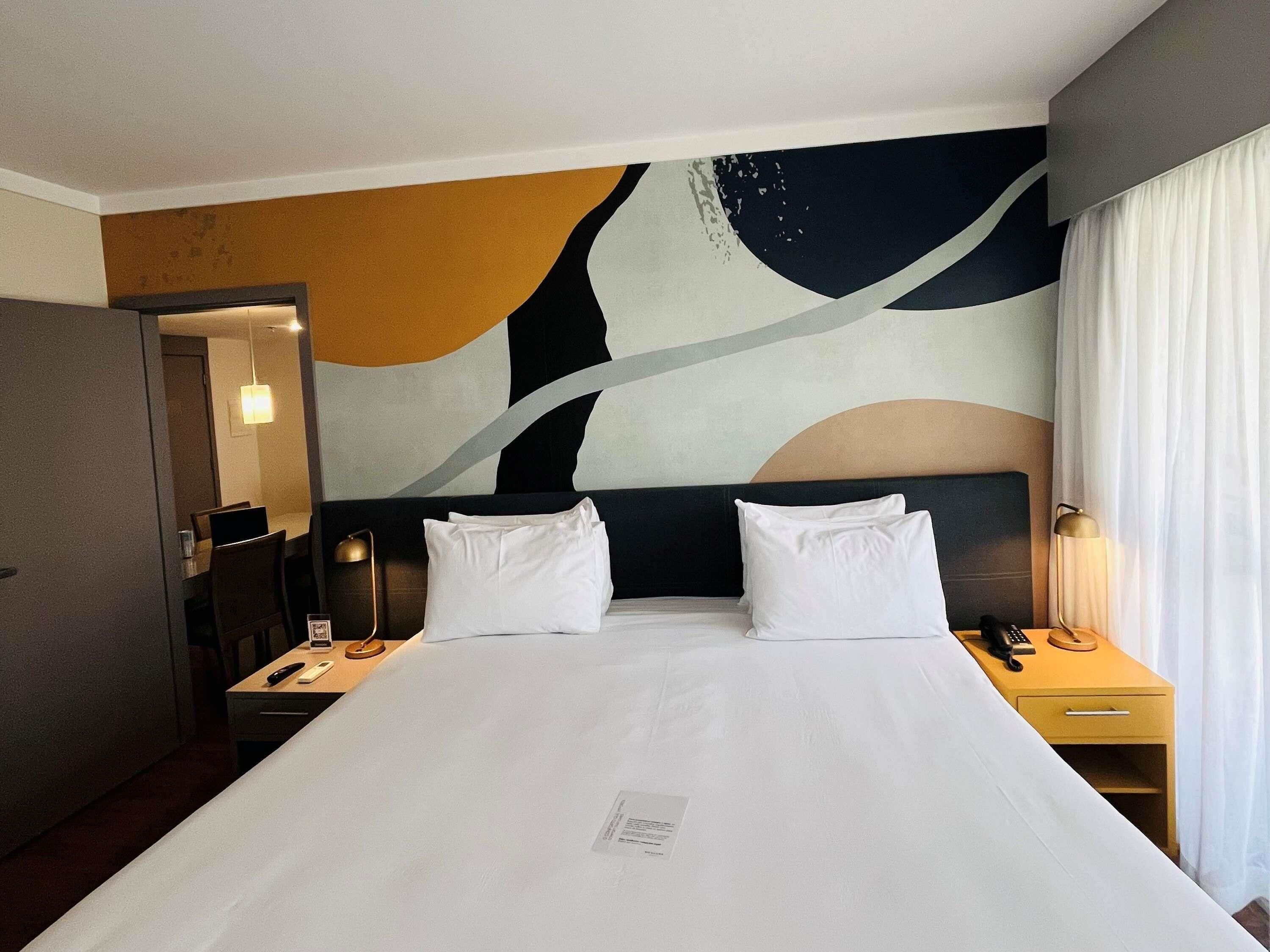 Habitación de hotel con una cama grande, dos mesitas de noche, un escritorio y una pared decorada con arte abstracto.