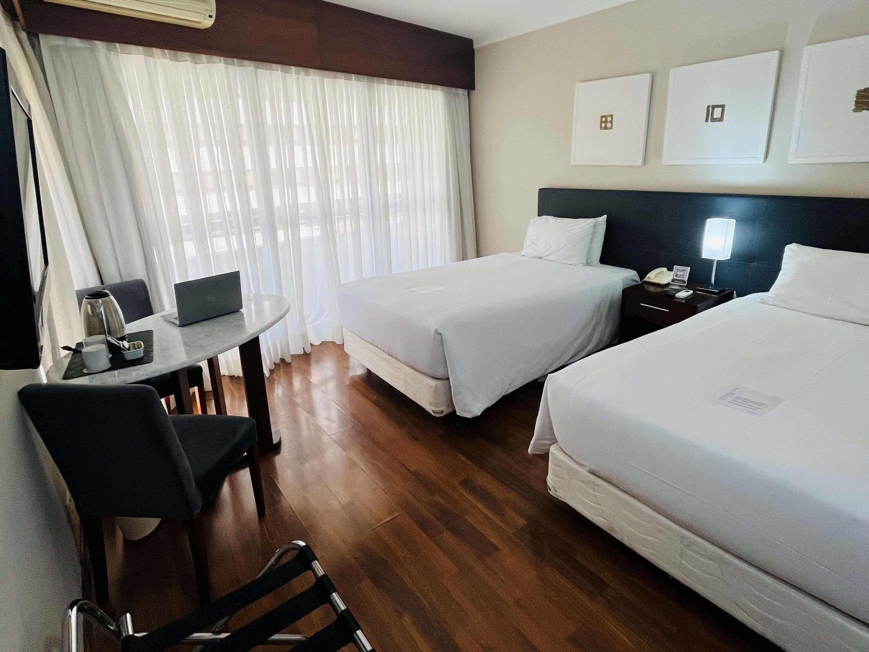 Habitación de hotel con dos camas, un escritorio, una silla, una computadora portátil, una cafetera y una mesita de noche con lámpara.