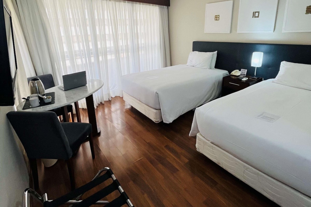 A hotel room with two beds, a desk, a chair, a laptop, a coffee maker, and a nightstand with a lamp.