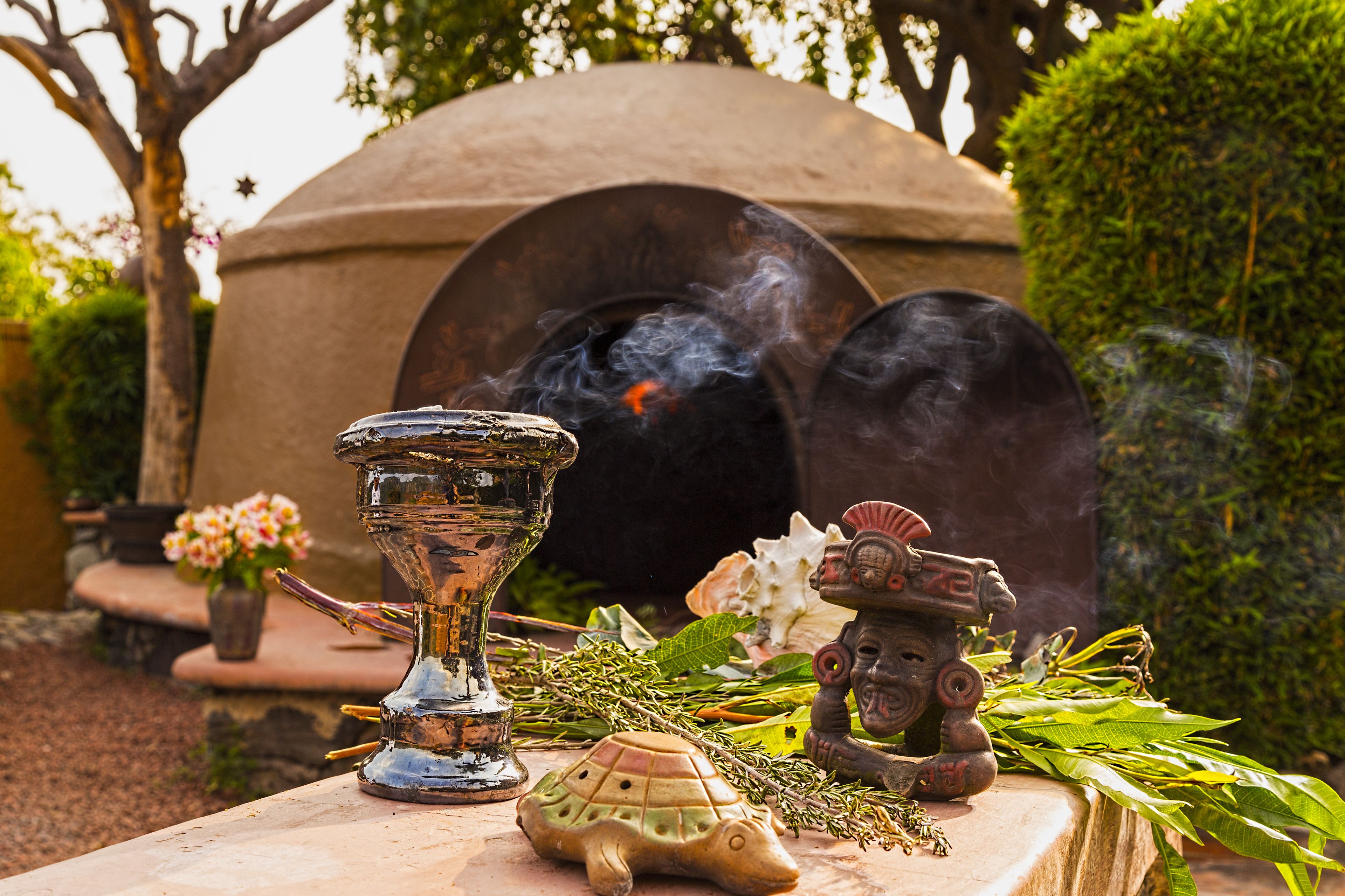 Una estructura de piedra con humo, una tortuga de cerámica y una pequeña estatua sobre una mesa con follaje y flores.