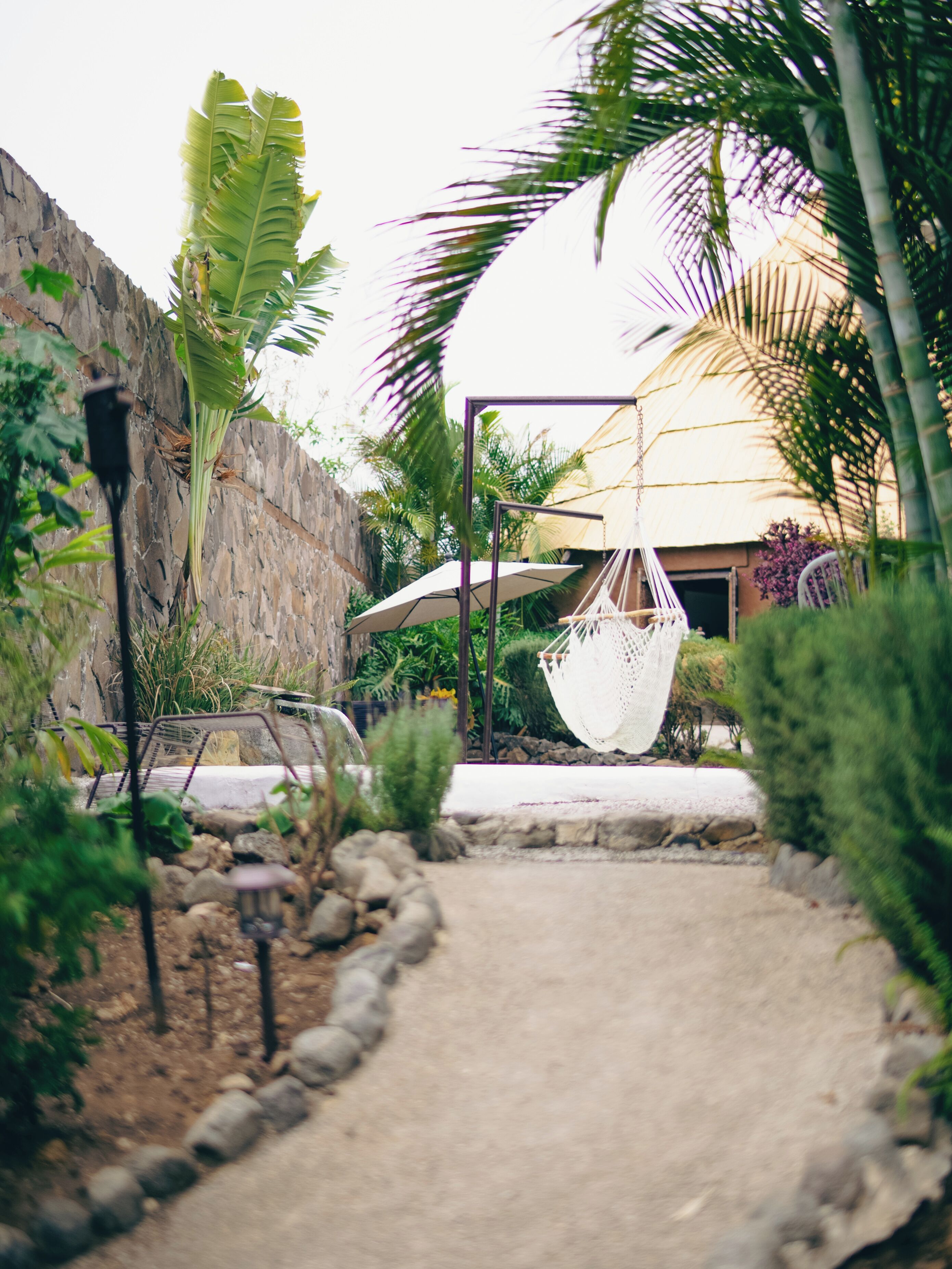Un sentier mène à un fauteuil suspendu et à un hamac dans un jardin agrémenté d’un mur de pierre et de plantes tropicales.