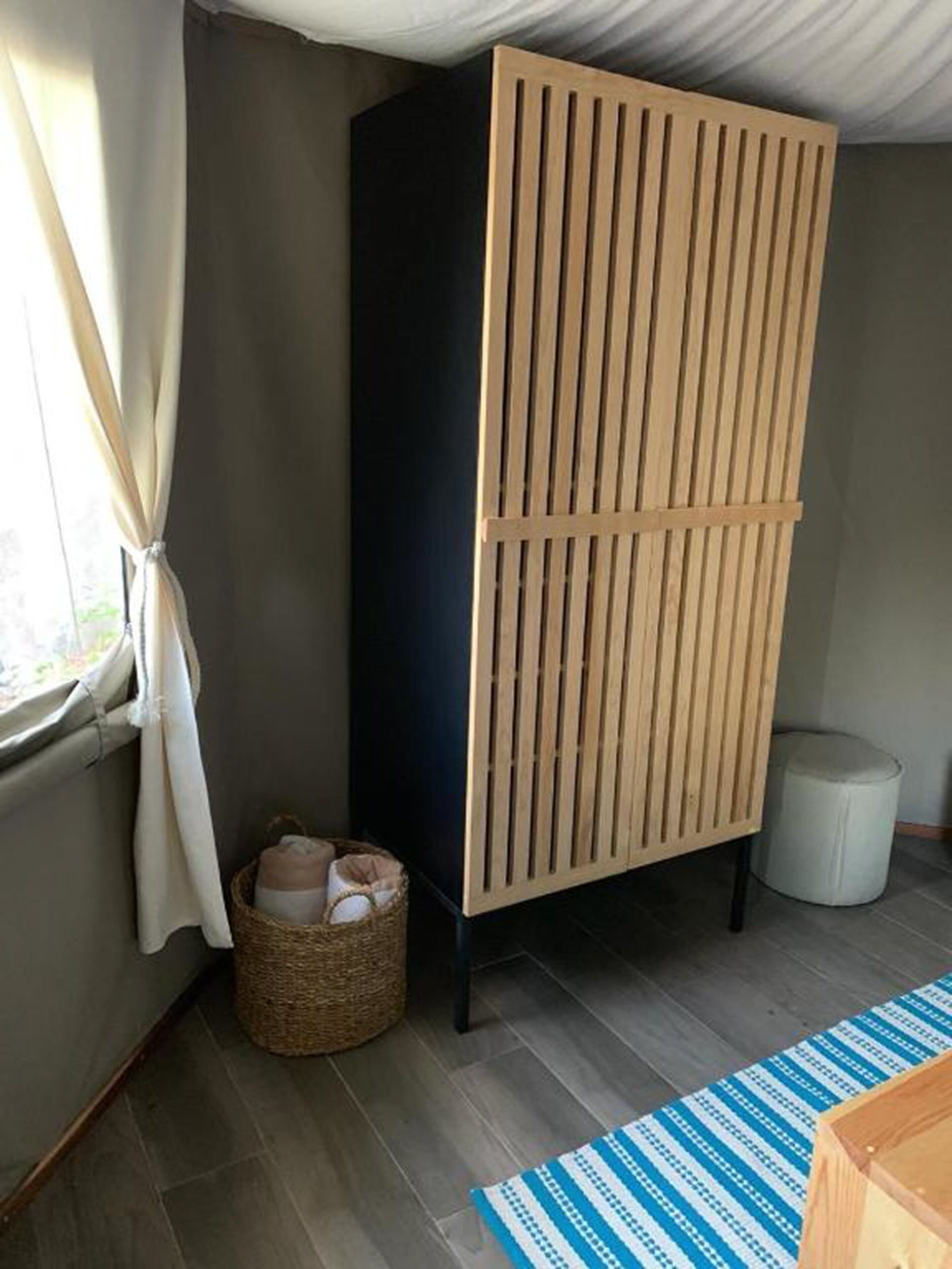 Une pièce comprenant une grande armoire de couleur sombre, un panier en osier contenant divers objets, un tapis à rayures bleues et blanches, et une poubelle blanche.