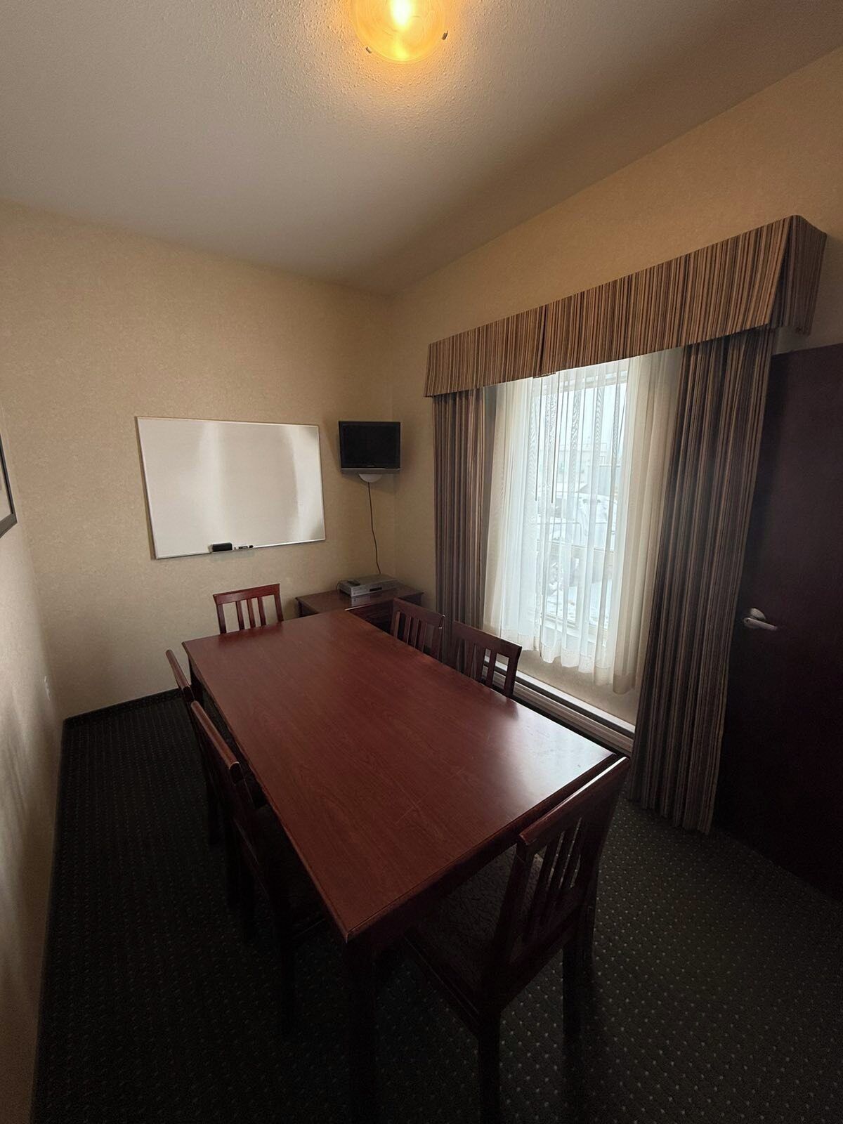 A conference room with a wooden table, chairs, a whiteboard, a television, and a window with curtains.