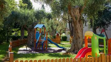 Children can play on a slide, climbing frame, and swing set in a shaded area surrounded by lush green grass and mature trees.