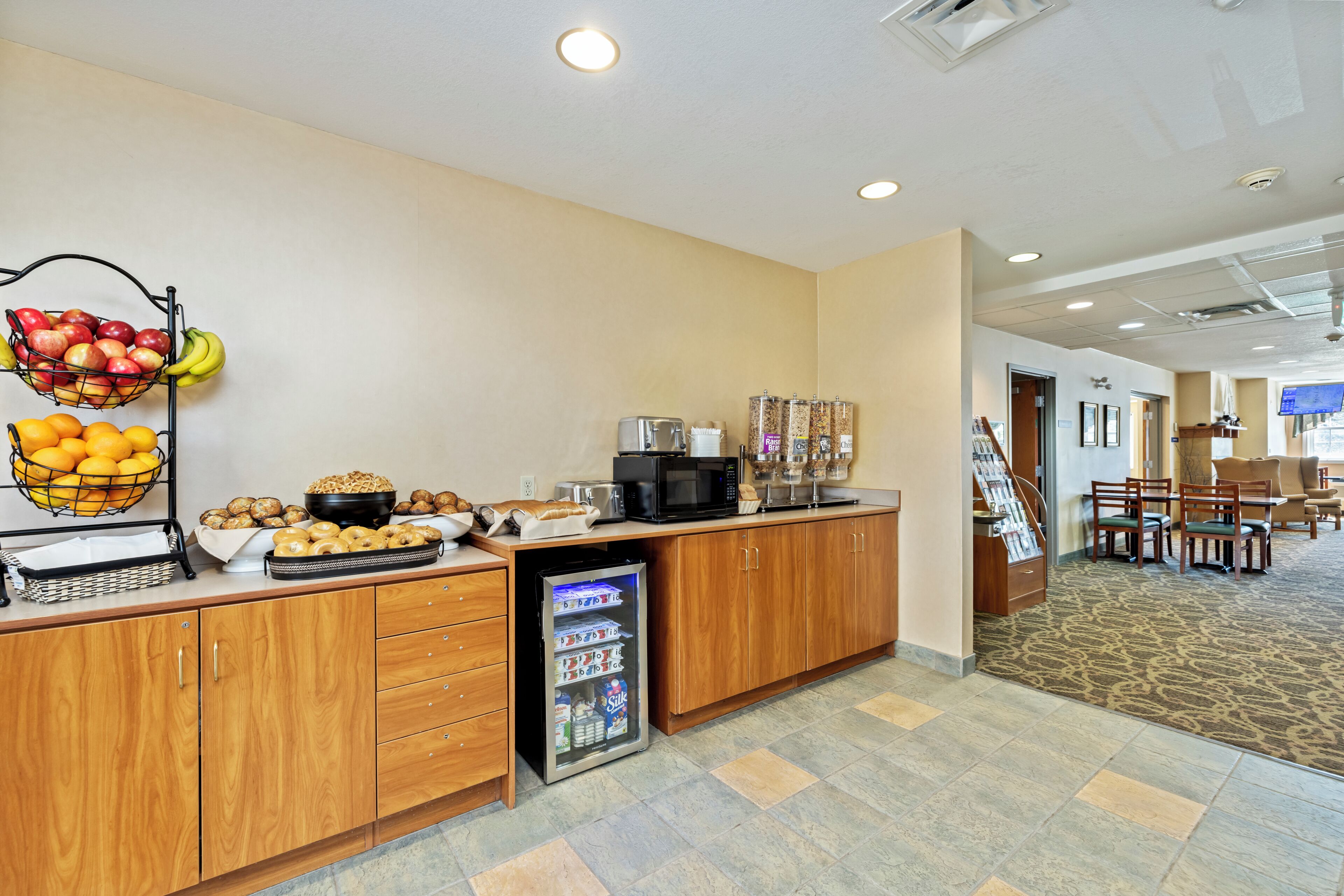 Een hotel receptie met een fruitmand, een koffieapparaat, een koelkast, en een zithoek met tafels en stoelen.
