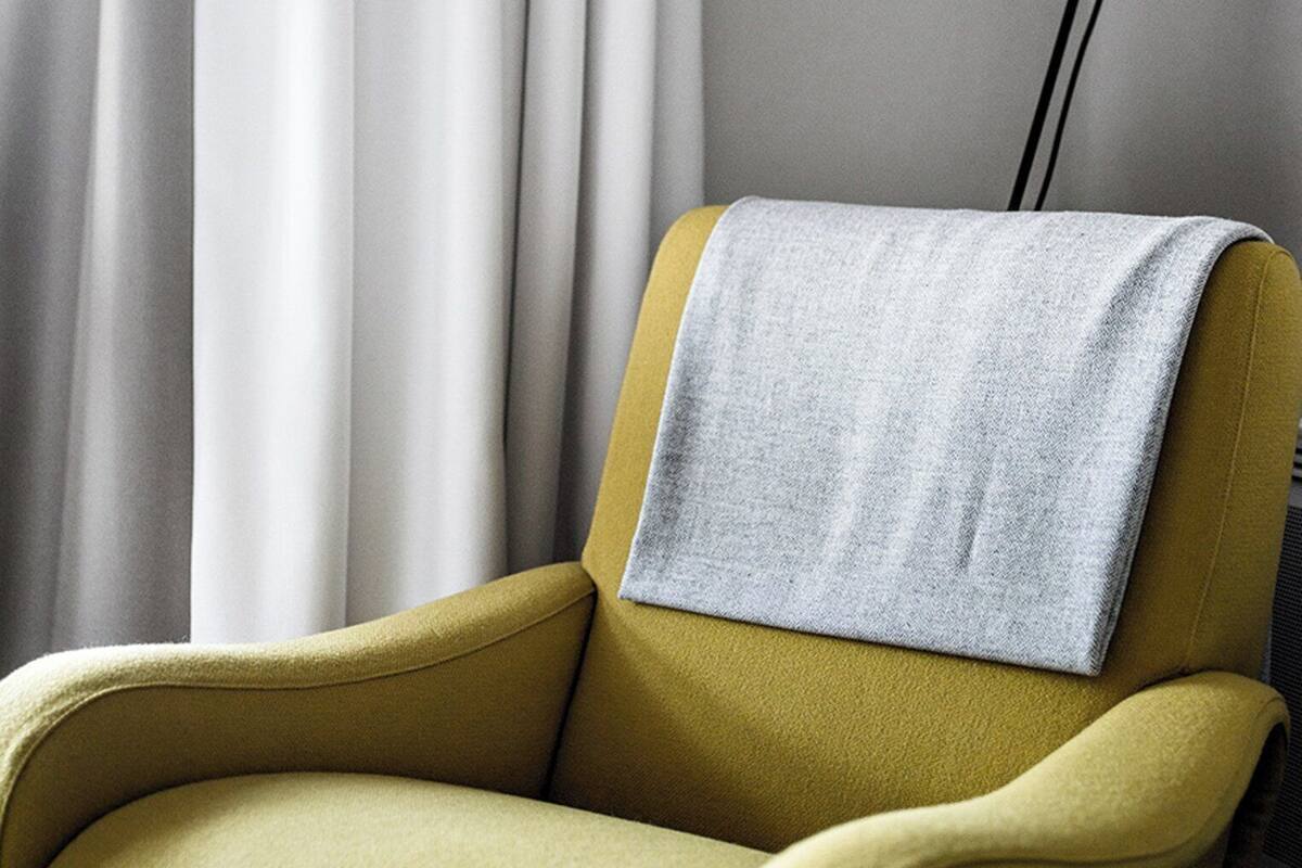 Un sillón amarillo con una manta gris, una lámpara de pie y un juego de cortinas blancas.