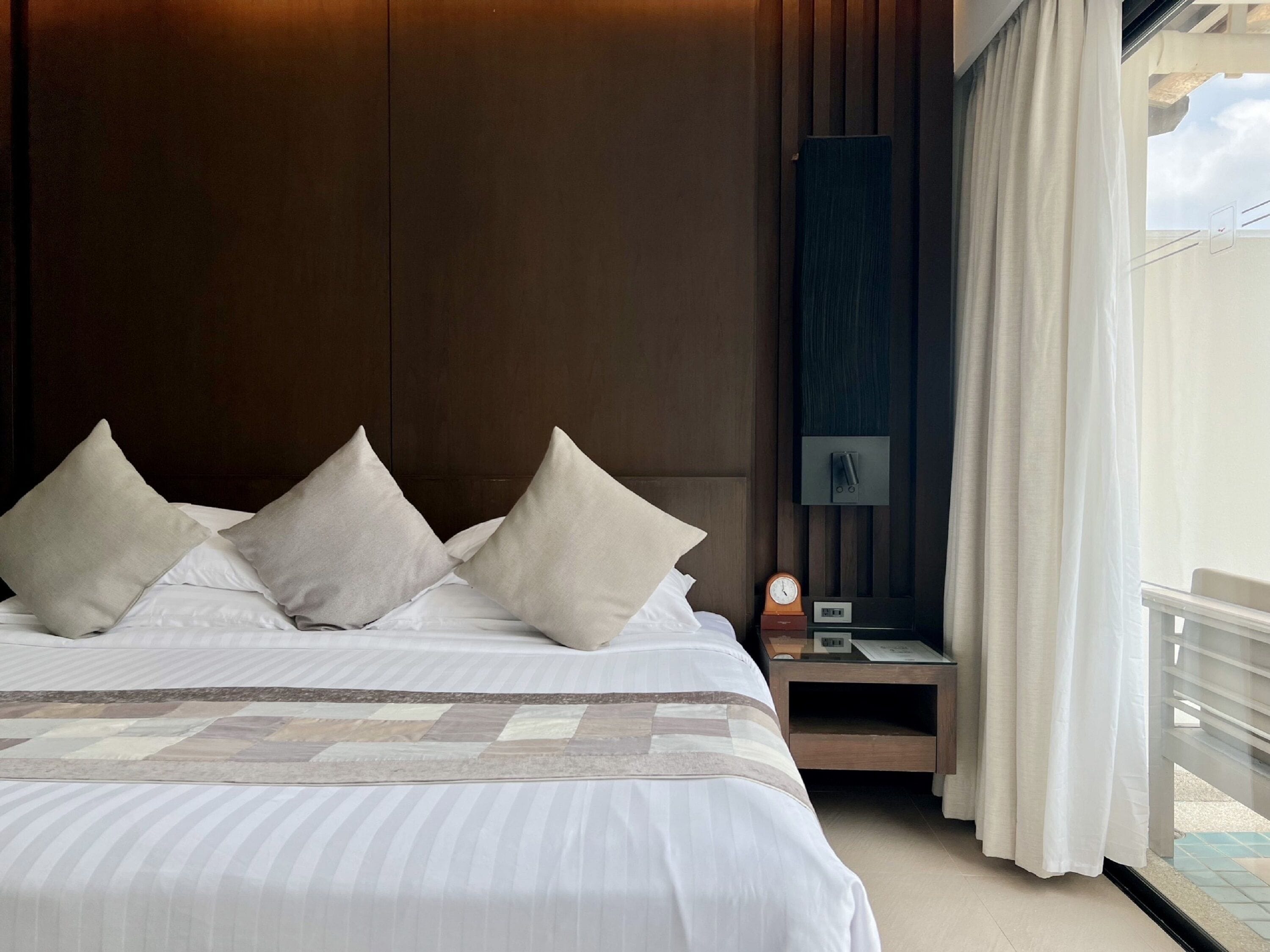 A neatly made bed with three pillows, a bedside table with a clock and a small lamp, and a view of the outdoors through a window with curtains.