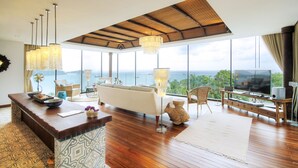 A spacious living room with a large window overlooking the sea, a wooden coffee table, a sofa, and a TV on a wooden stand. - Cape Panwa Hotel (Wichit)
