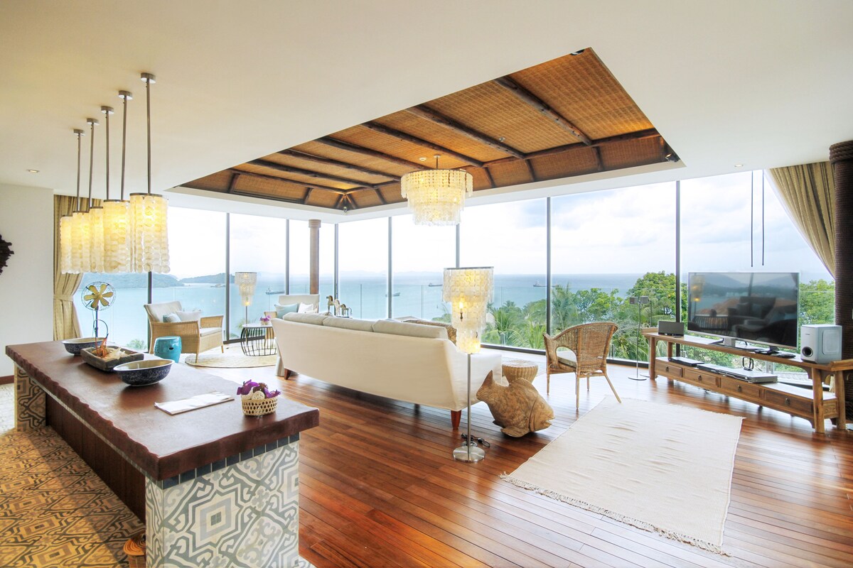 A spacious living room with a large window overlooking the sea, a wooden coffee table, a sofa, and a TV on a wooden stand.