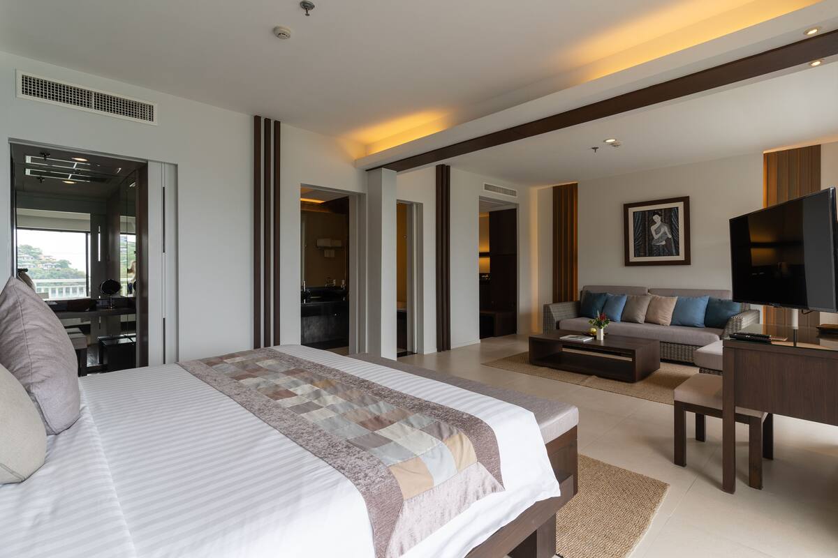 A modern hotel room with a large bed, a sofa, a TV, and a desk. There are multiple doors leading to other rooms and a balcony.