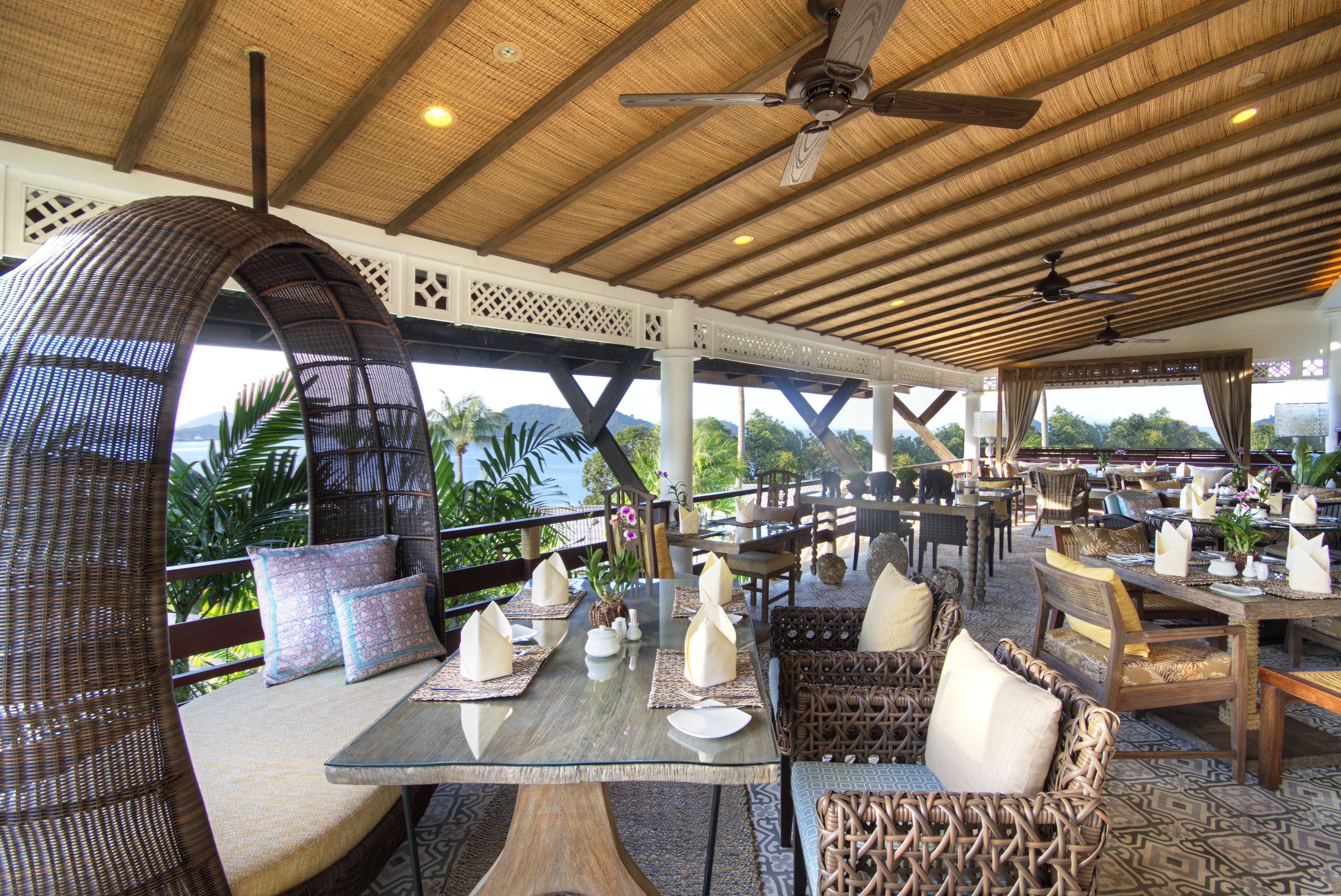 An outdoor dining area with wicker furniture, tables set with plates and napkins, and a view of the ocean.