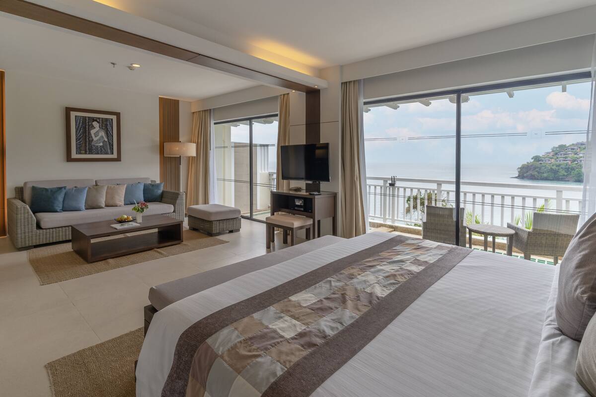 A modern hotel room with a large bed, a sofa, a TV, and a balcony with a view of the sea.