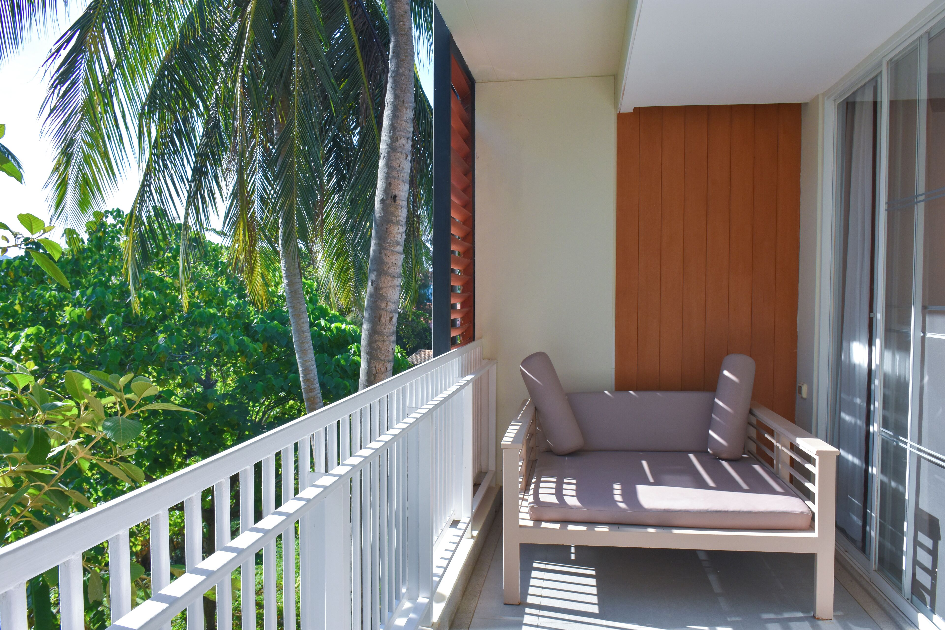 A balcony with a white railing, a cushioned bench, and a view of lush greenery and palm trees.
