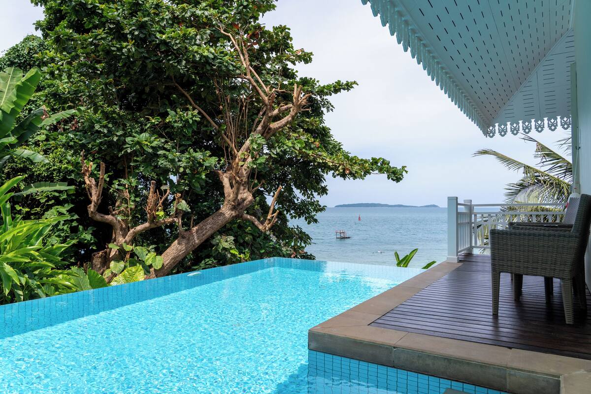 A pool with a view of the sea, surrounded by lush greenery and a wooden deck with a chair.