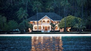 A large, two-story building with a balcony, surrounded by palm trees and a beach.
