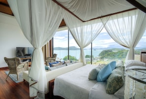 A bedroom with a large bed, a television, wicker furniture, and a view of the sea. - Cape Panwa Hotel (Wichit)