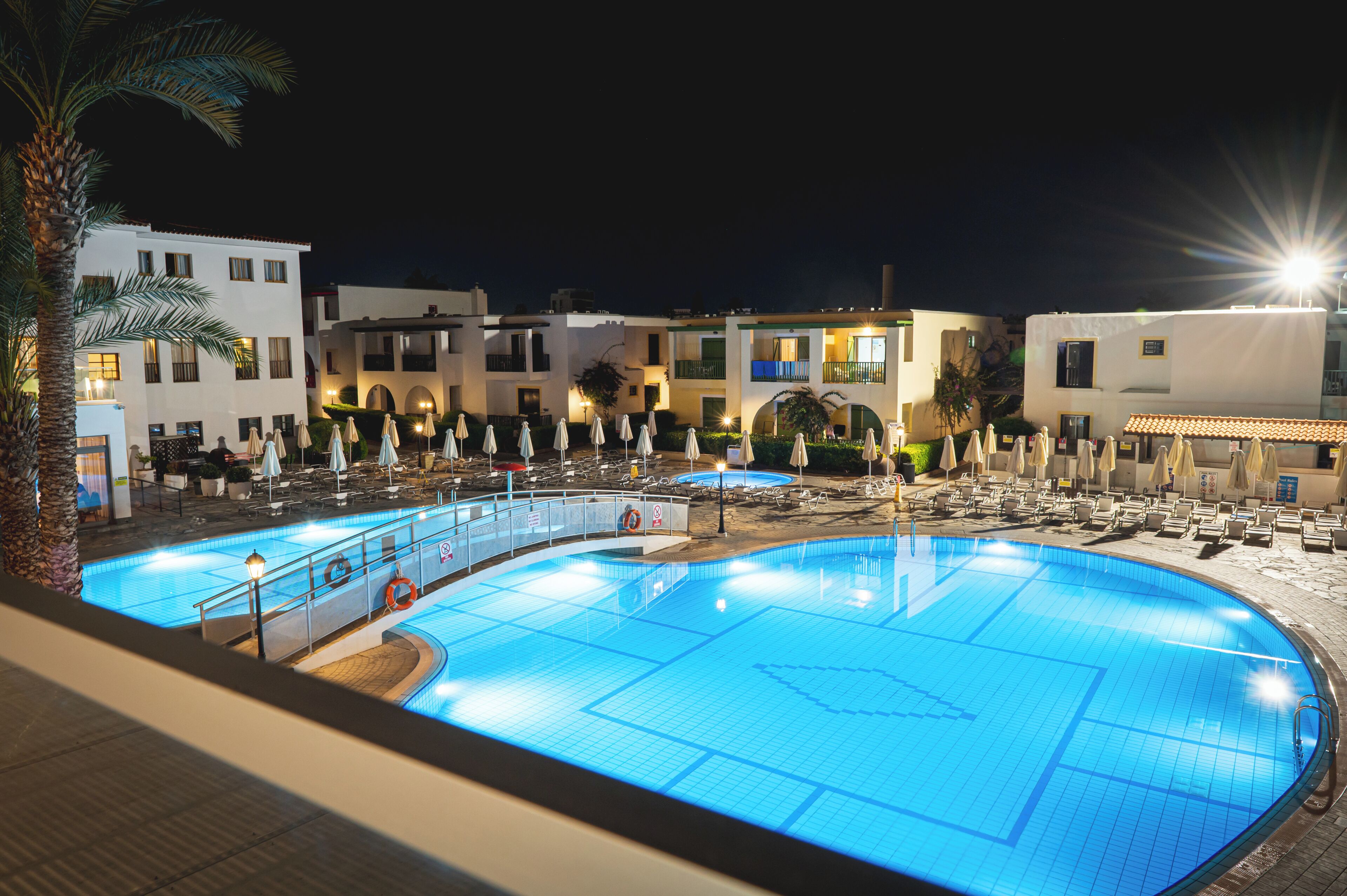 Une piscine dotée d’une terrasse carrelée, entourée de chaises longues et de parasols, avec des bâtiments en arrière-plan.
