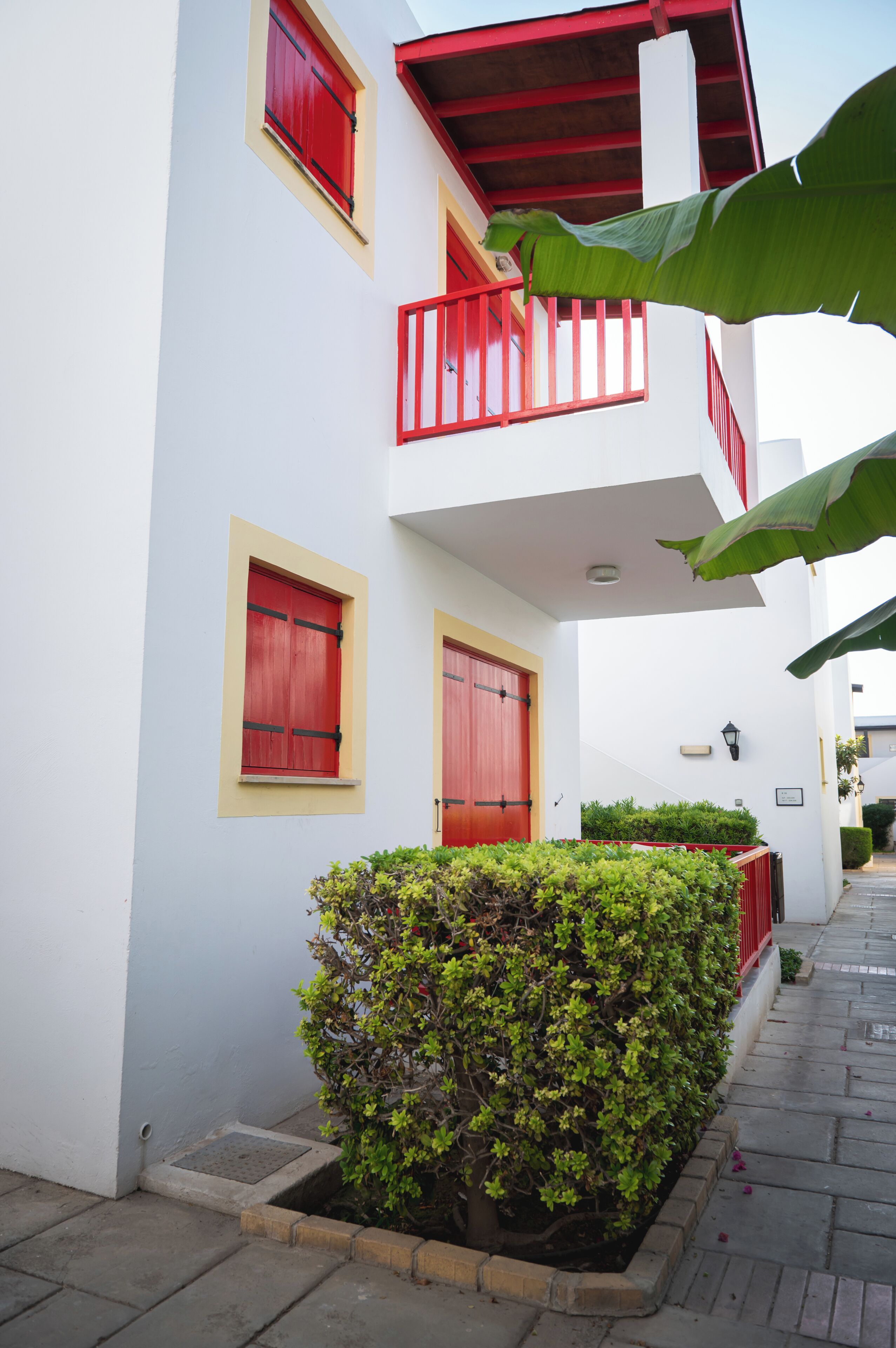 A building with red-framed windows and doors, a small balcony with a red railing, and a well-maintained hedge in front.