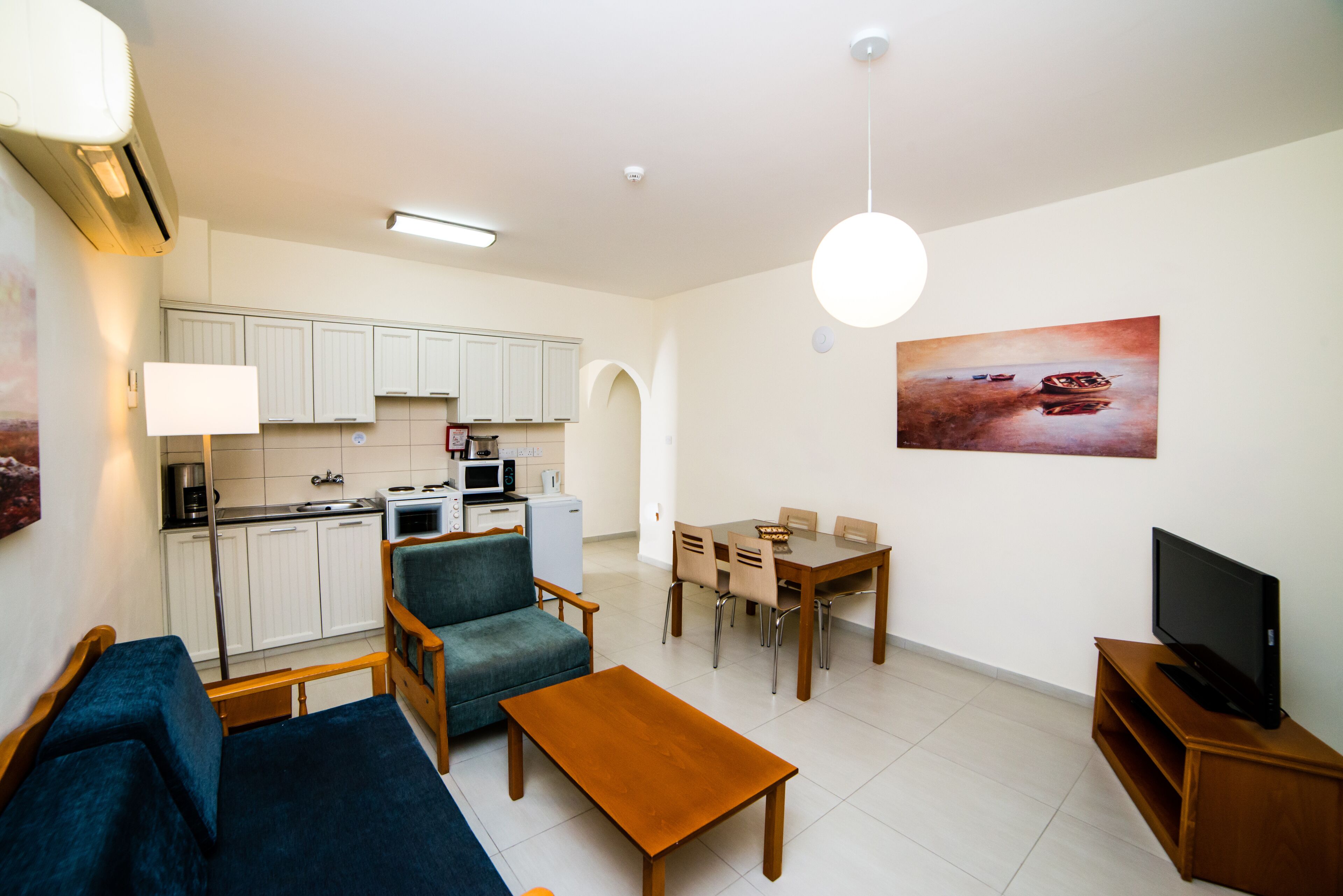 A modern living room with a sofa, coffee table, dining table, and chairs. A TV is on a wooden stand. The kitchen is visible with white cabinets and appliances.