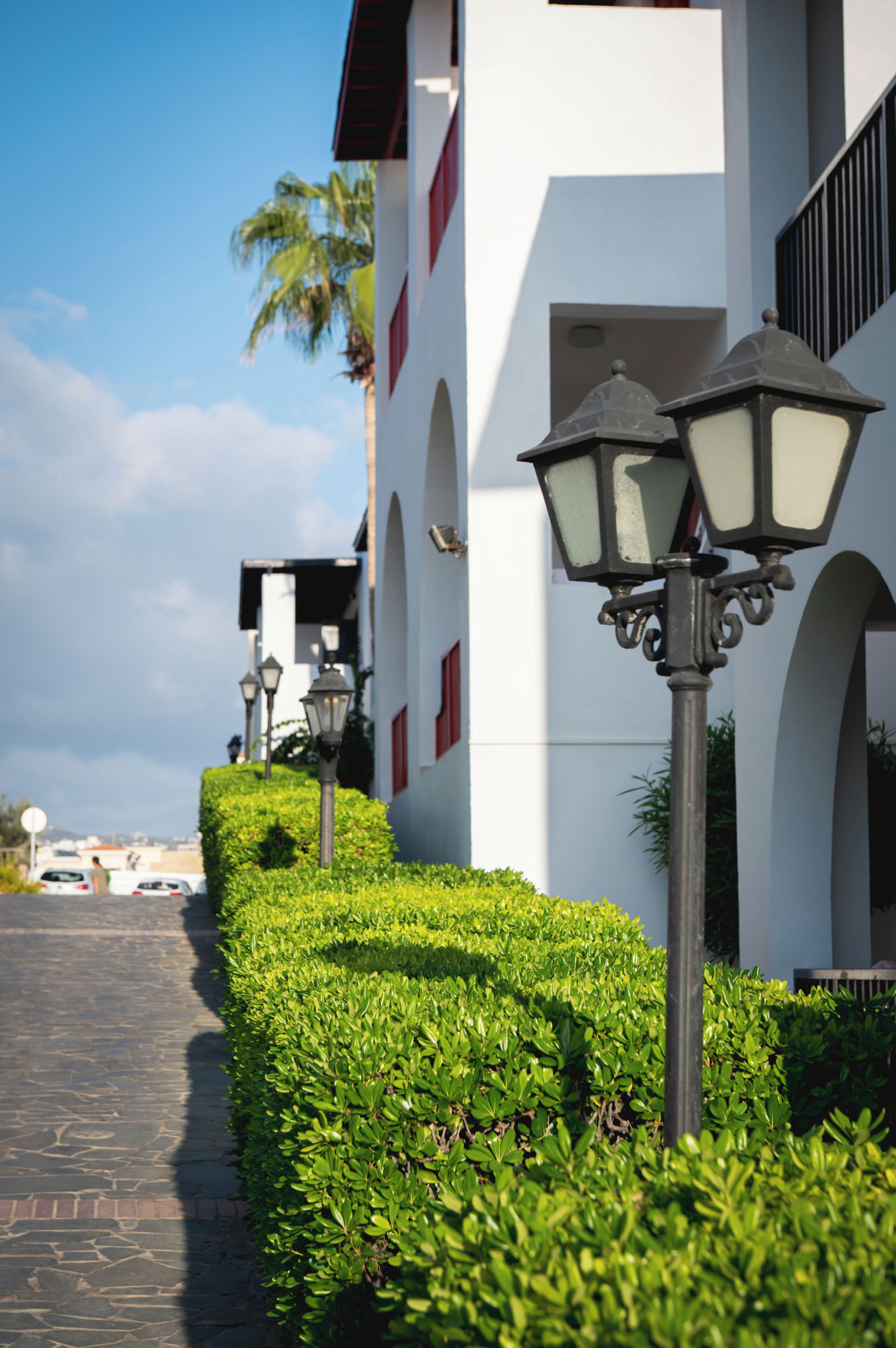 A pathway lined with neatly trimmed hedges and two street lamps, leading to a building with a white facade and red accents.