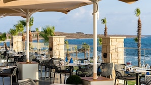 An outdoor dining area with tables and chairs, overlooking a coastal landscape with palm trees and a view of the sea. - Kefalos Beach Tourist Village (Paphos)