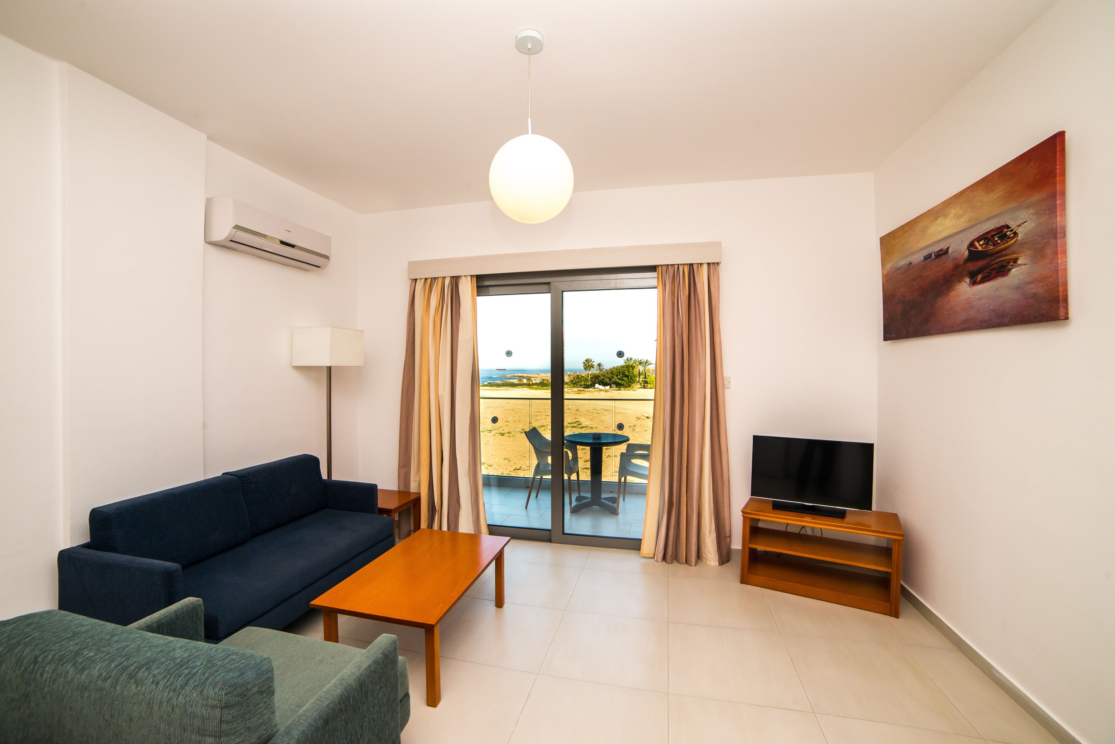 A modern living room with a sofa, a coffee table, a TV on a wooden stand, and a balcony with a view.