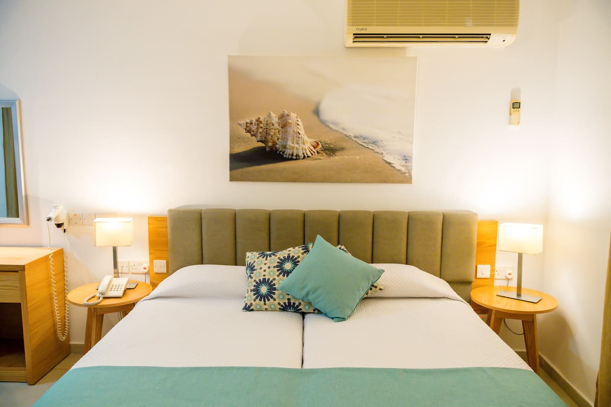 Une chambre d’hôtel avec un lit, deux tables de chevet, un climatiseur fixé au mur et un tableau représentant un coquillage accroché au mur.