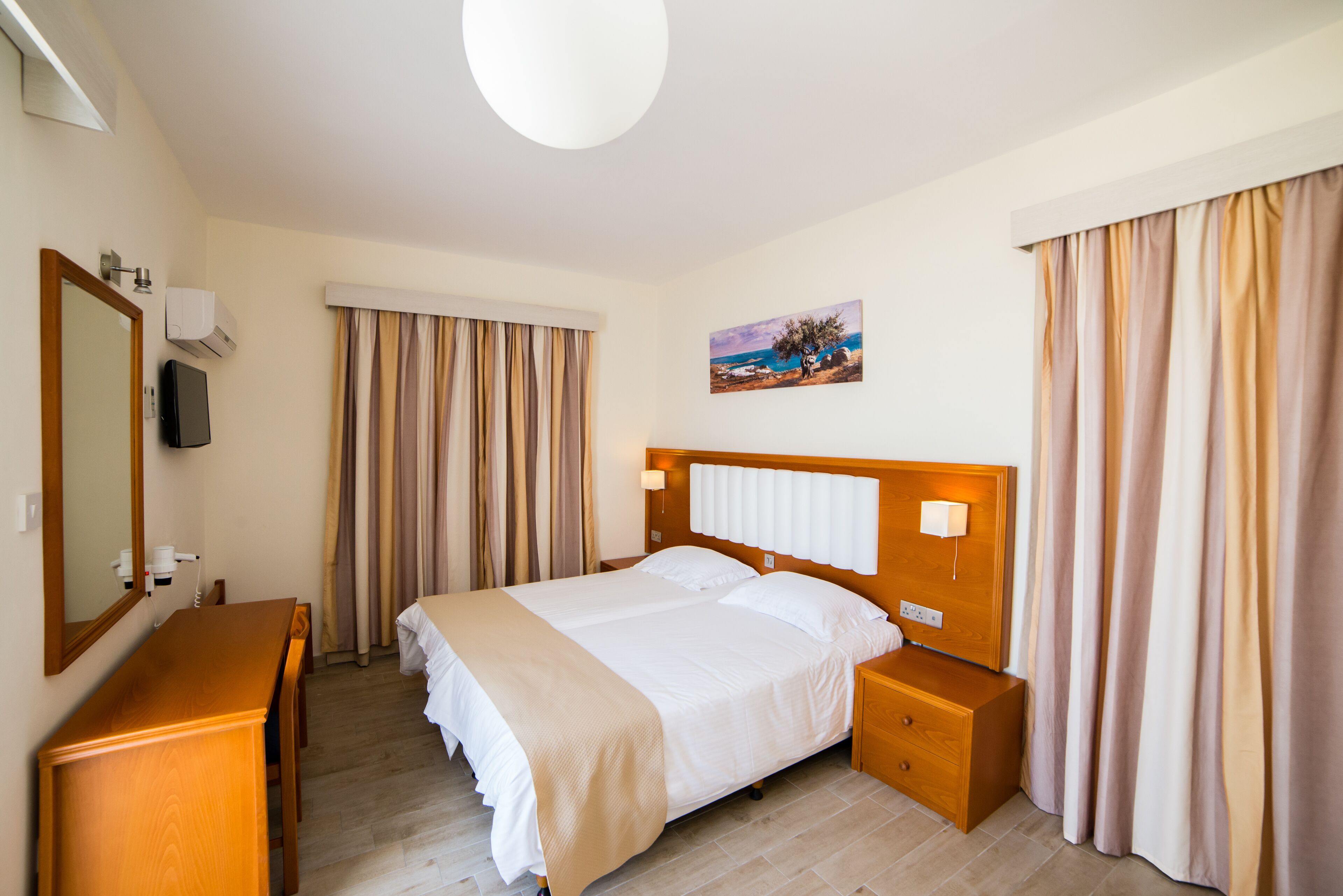 A hotel room with a bed, a wooden nightstand, a desk, a mirror, and a painting on the wall.