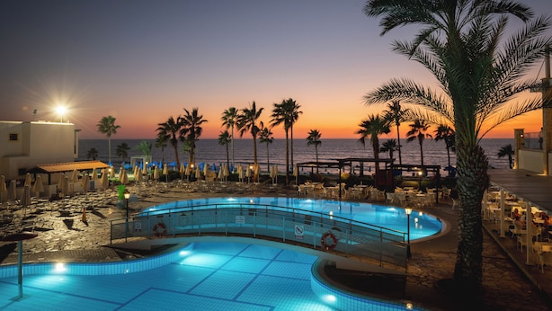 A pool area with a curved design, palm trees, beach chairs, and umbrellas. - Kefalos Beach Tourist Village (Paphos)