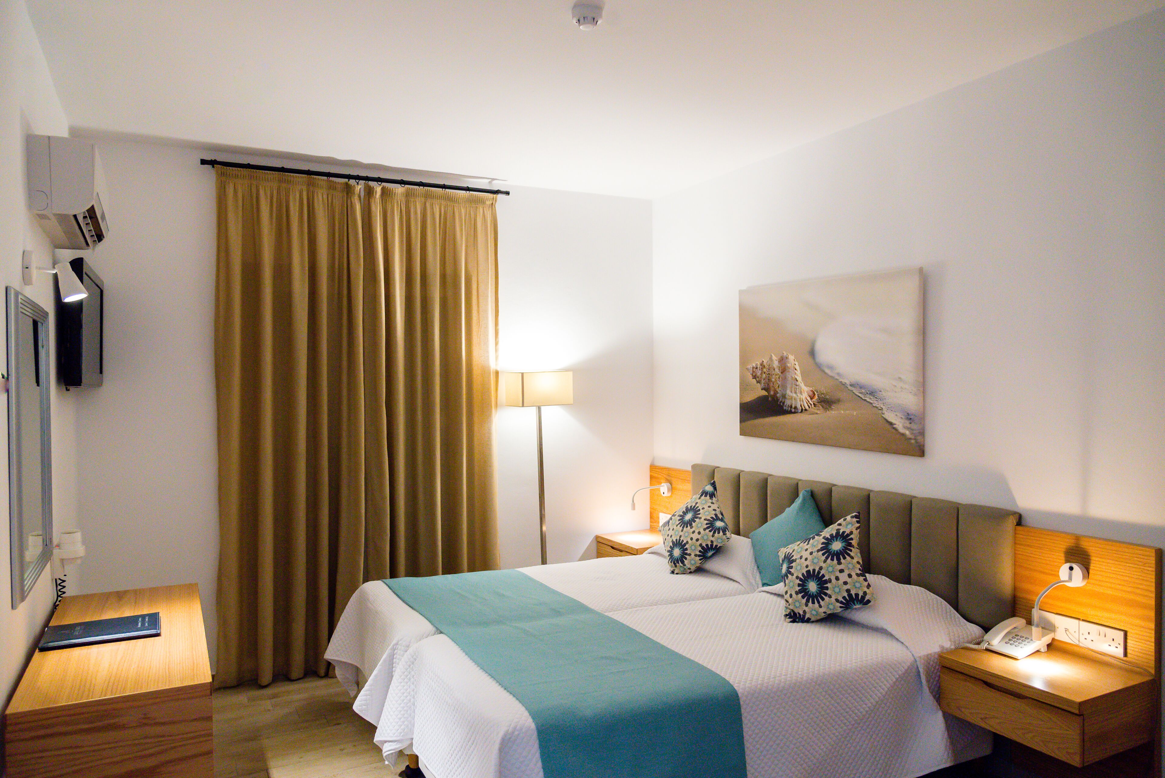 Ein Hotelzimmer mit Bett, Nachttisch, Lampe, einem Gemälde und einem Vorhang.