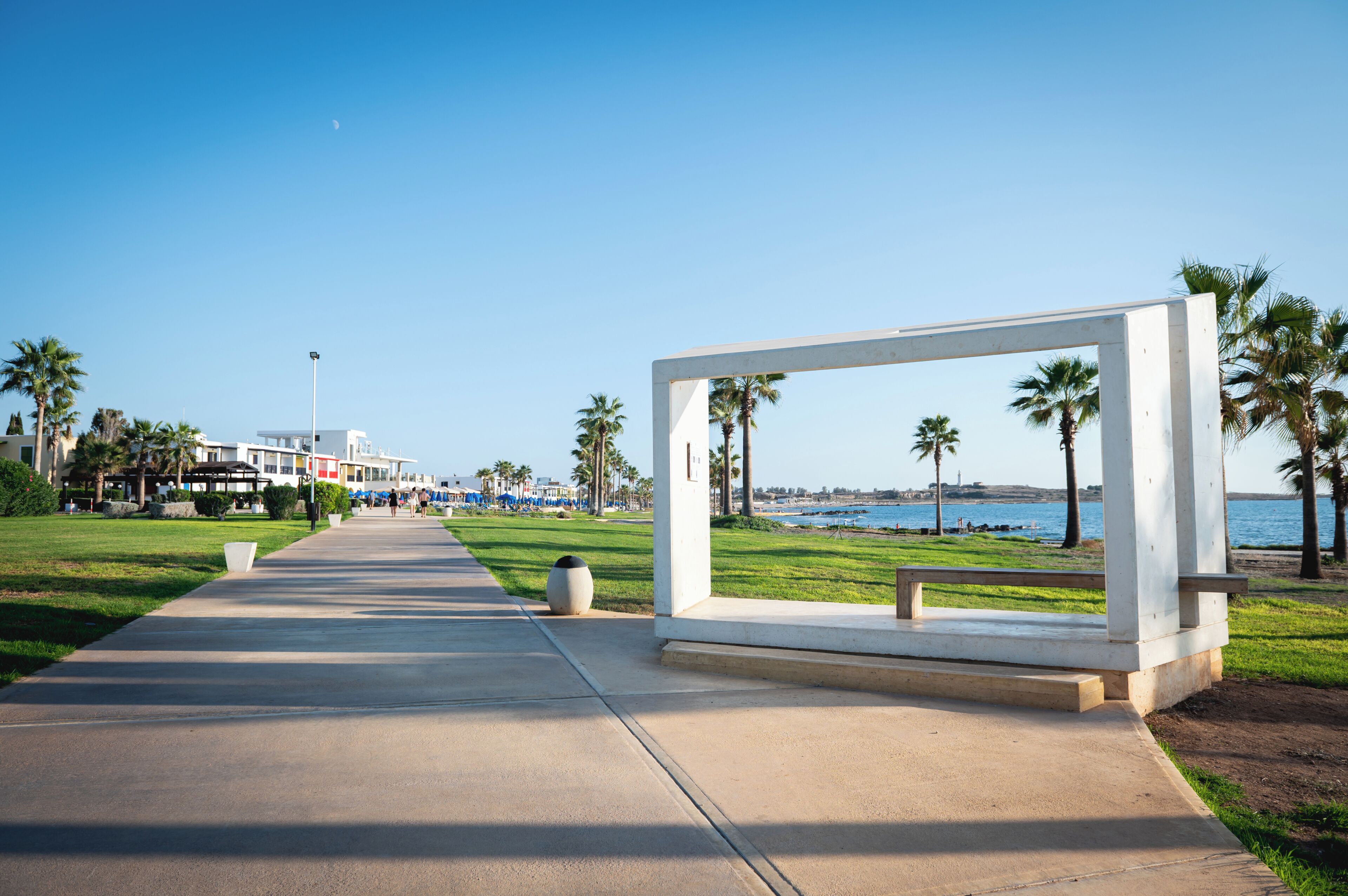 A pathway leading to a white structure with a bench, palm trees, and a view of the ocean.
