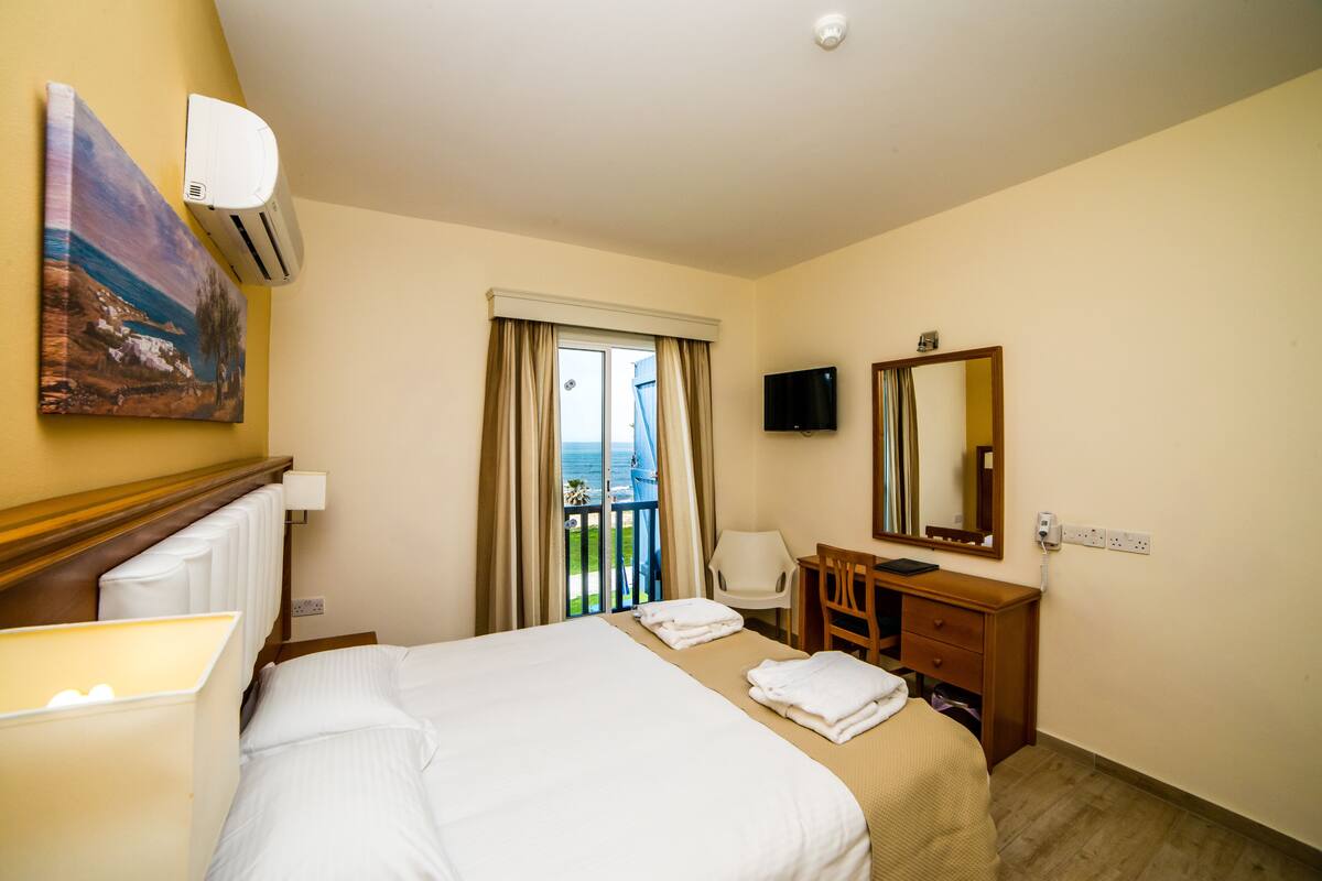 Une chambre d’hôtel équipée d’un lit, d’un bureau, d’une chaise, d’une télévision, d’un miroir et d’une fenêtre donnant sur l’extérieur.
