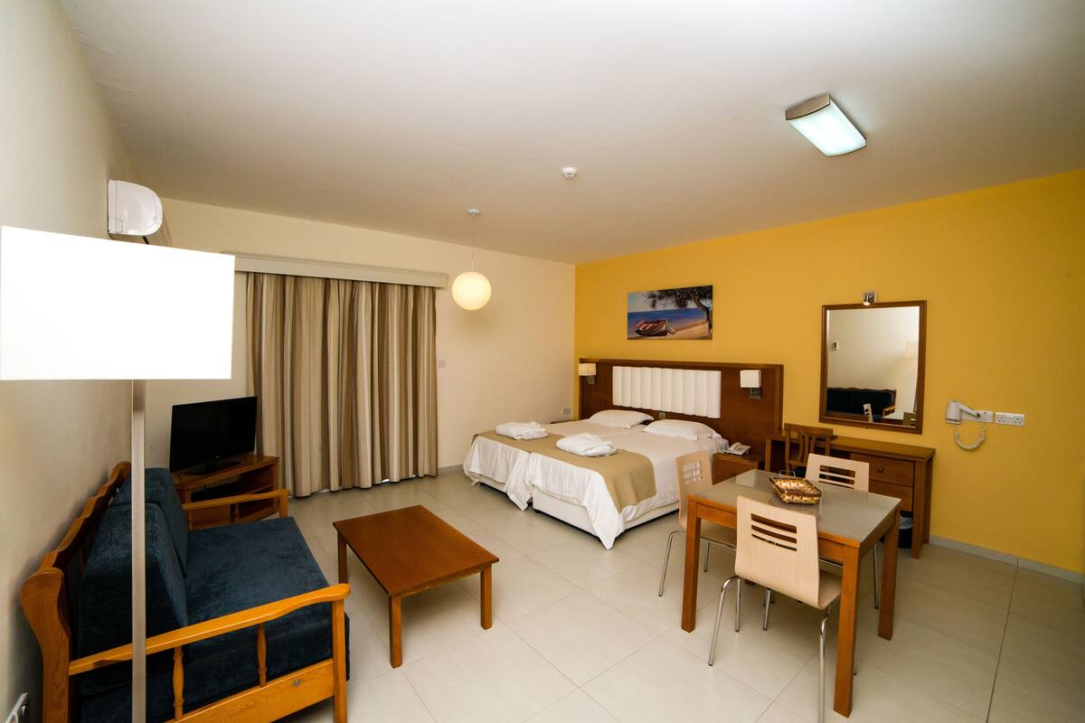 Une chambre d’hôtel avec un lit, un canapé, un bureau, une chaise, une télévision et une fenêtre avec des rideaux.