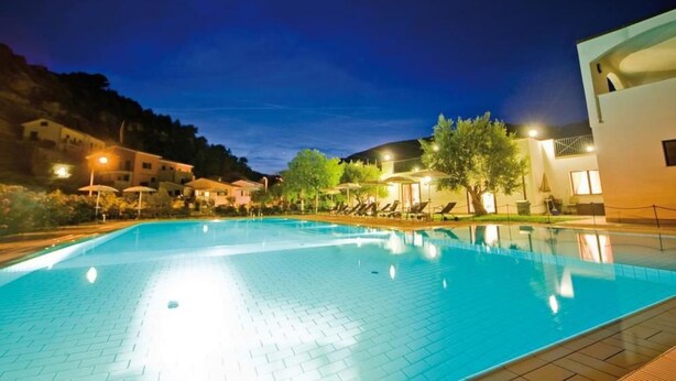 Una piscina con sillas de descanso y sombrillas, rodeada de edificios y árboles.