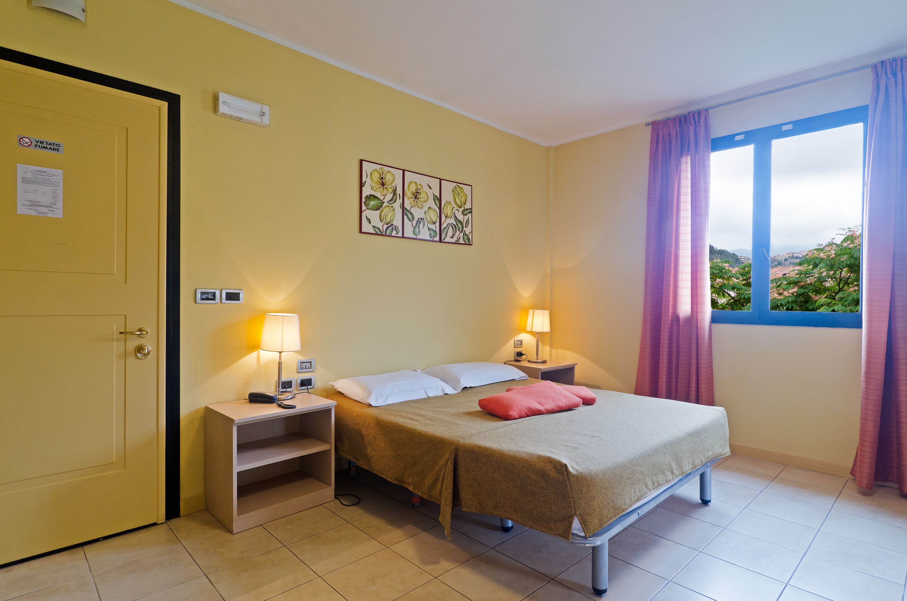 Ein Hotelzimmer mit Bett, Nachttischlampen, einem Nachttisch, einer Tür und einem Fenster mit Vorhängen.